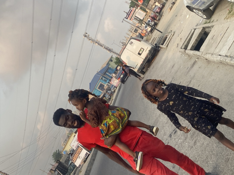 The image, taken in Badore, Nigeria, captures a man and two young girls outdoors on a street or roadside under an overcast sky. The photograph is tilted, giving a dynamic perspective to the scene.
In the foreground, a man with a beard, wearing a red t-shirt and red pants, smiles warmly as he carries a young girl. This girl, with dark braided or twisted hair, is dressed in a brown top and a vibrant, colorful patterned skirt. She looks intently towards the left side of the frame. To their right, a slightly older girl stands, looking towards the viewer or slightly past. She has dark hair with lighter streaks and is wearing a black long-sleeved dress adorned with gold heart-like patterns. Both girls appear to be young children.
The background reveals an urban or suburban setting. Power lines stretch across the grey, cloudy sky, and a tall communication tower stands prominently in the mid-ground. Various buildings line the street, including one with a distinctive blue facade. Vehicles, including a white van, are visible parked or driving further down the road. Concrete drainage covers are present along the road's edge. A sign in the distance is partially readable, displaying "BRODY ALPHA" and "LABORATORY," with "HOSPITAL" also partly visible. The overall scene depicts a casual daytime outing for the man and the children, under a diffused, cloudy light. The man's smiling expression suggests a pleasant moment.