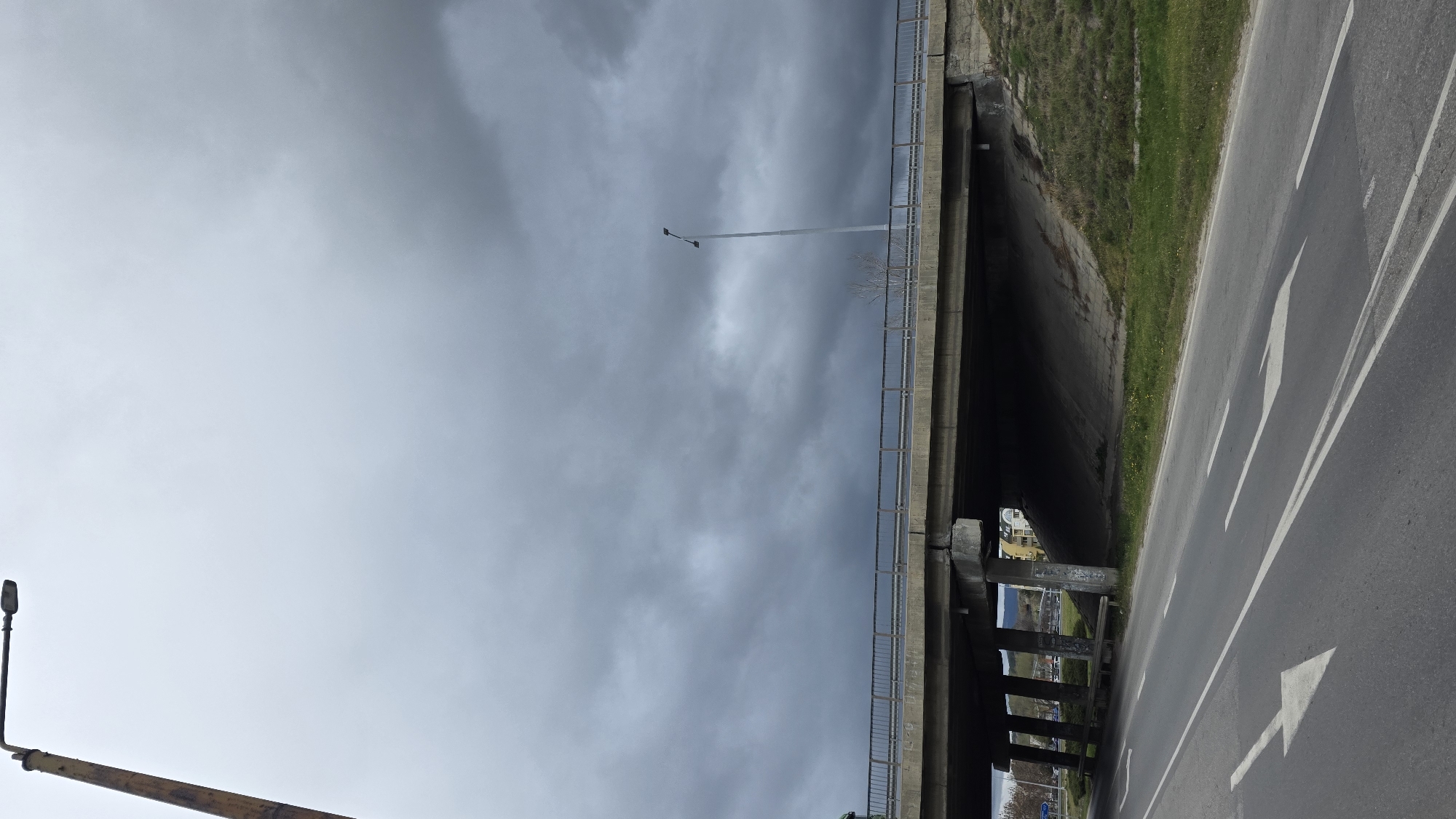 The image depicts an overcast day at a road in Blagoevgrad, Bulgaria. The road runs beside and under a concrete overpass or bridge. Arrow markings are painted on the asphalt. Vegetation grows on the embankment next to the road. A streetlight stands on the road, while a taller lamppost is visible on the overpass. Through the overpass, there is a glimpse of buildings and structures in the distance.