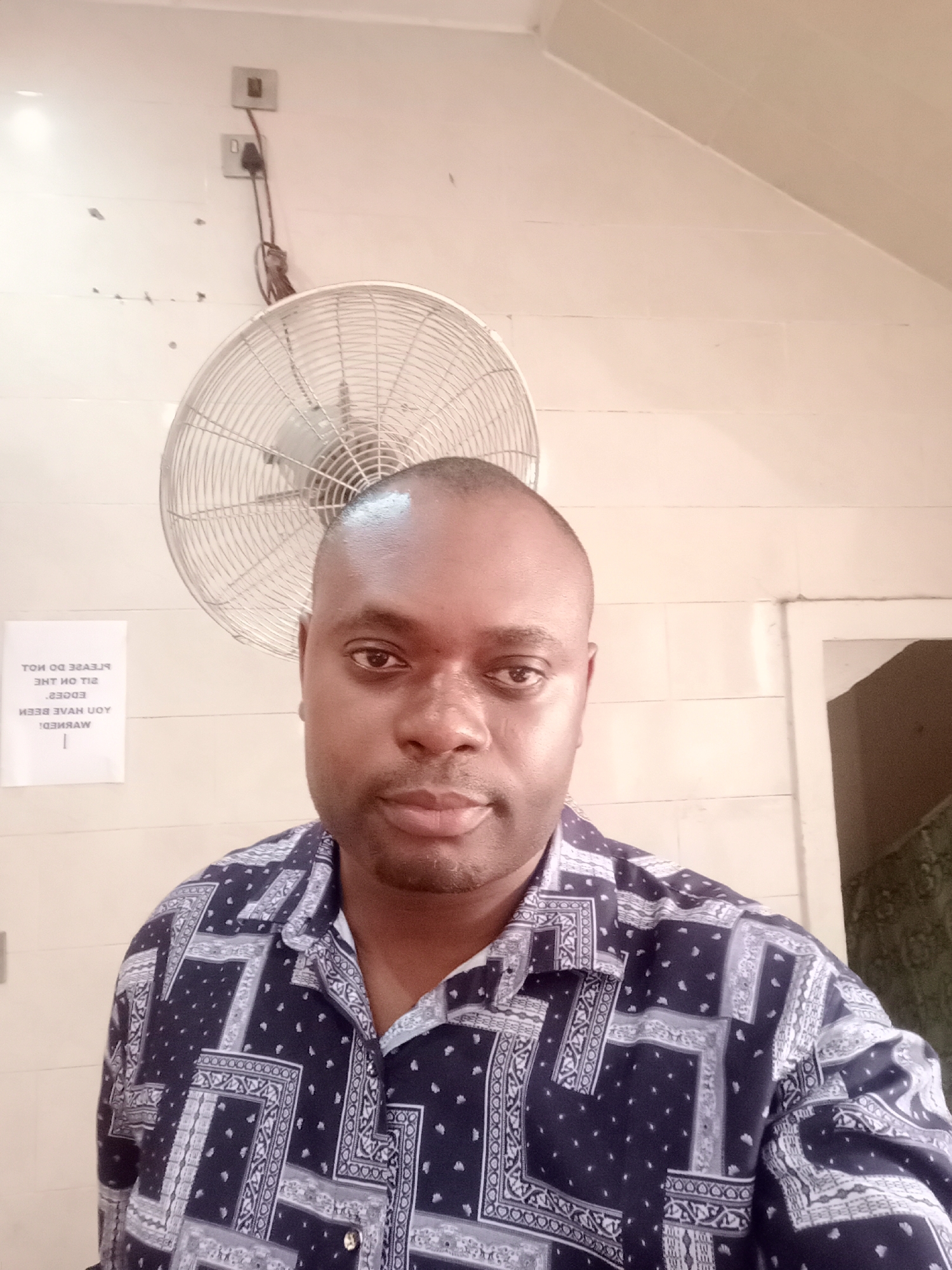 An adult man, with dark skin and a shaved head, is captured in a close-up, eye-level shot, looking directly at the camera. He wears a dark blue collared shirt with a white and light blue geometric, paisley-like pattern. His expression is neutral to slightly serious.
The man fills much of the frame, with a simple indoor setting visible behind him. A wall-mounted, industrial-style oscillating fan with a silver metal cage is positioned to his upper left, partially obscured by his head. The fan appears stationary. Above and to the right of the fan, a couple of grey electrical outlets or switches are visible on the plain, light-colored wall, with dark cables hanging from them.
On the wall to the man's left, a white paper sign is taped, displaying inverted text. Upon correction, the text reads: "PLEASE DO NOT SIT ON THE EDGES. YOU HAVE BEEN WARNED!" To the far right, a glimpse of an open doorway or passage with darker interior is visible. The bright, even lighting suggests it is daytime. The presence of the fan indicates a warm climate, consistent with the specified location of Gwarinpa, Nigeria. The scene appears to be an everyday, functional indoor environment.