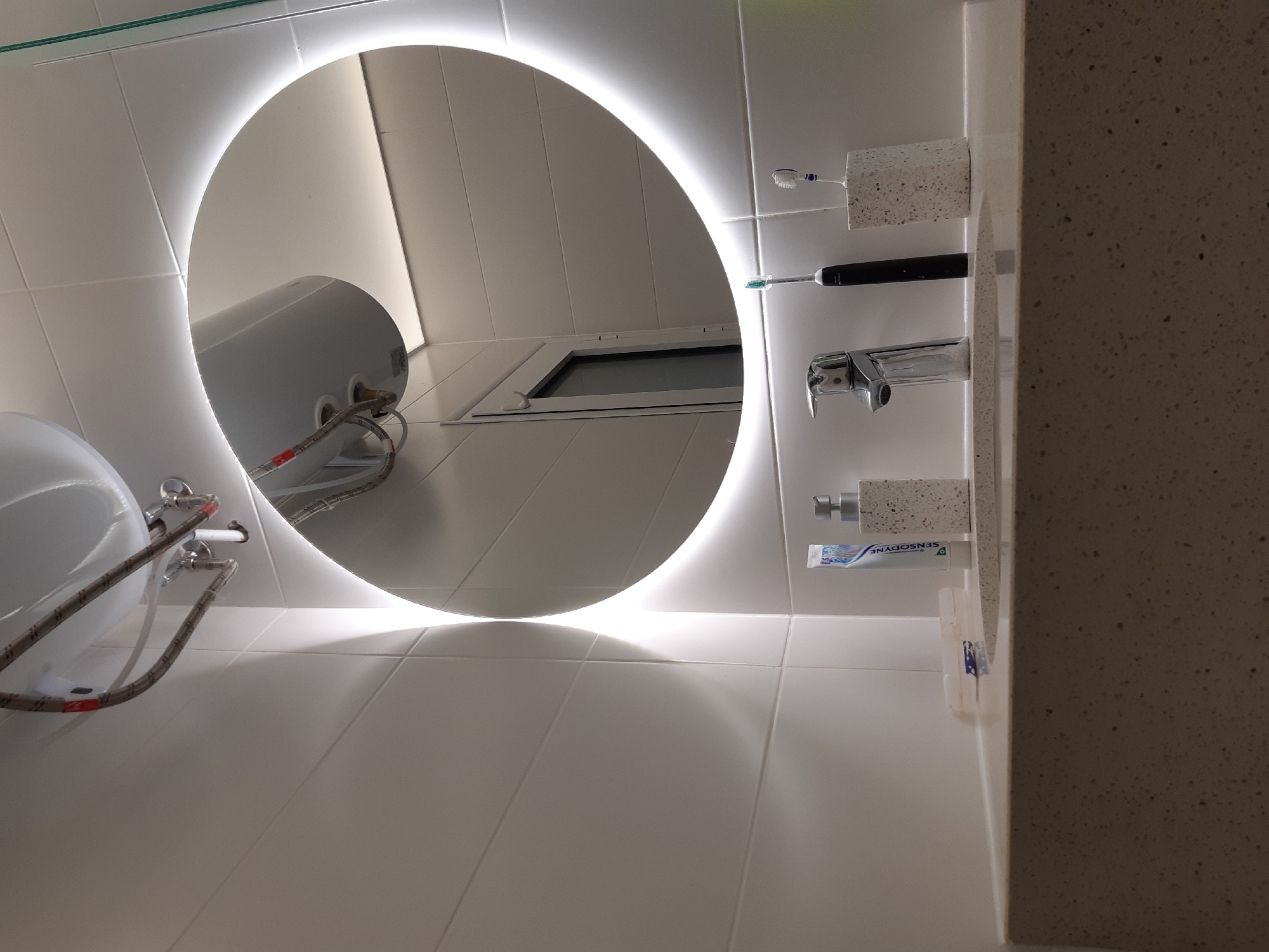 The image shows a bathroom interior. A round, illuminated mirror reflects the scene. Visible in the mirror are white tiled walls, a water heater, and a window. To the right of the mirror is a sink with a spotted countertop. Toothbrushes, a tube of toothpaste, and a faucet are attached to the wall. The location is likely a bathroom in Plovdiv, Bulgaria.