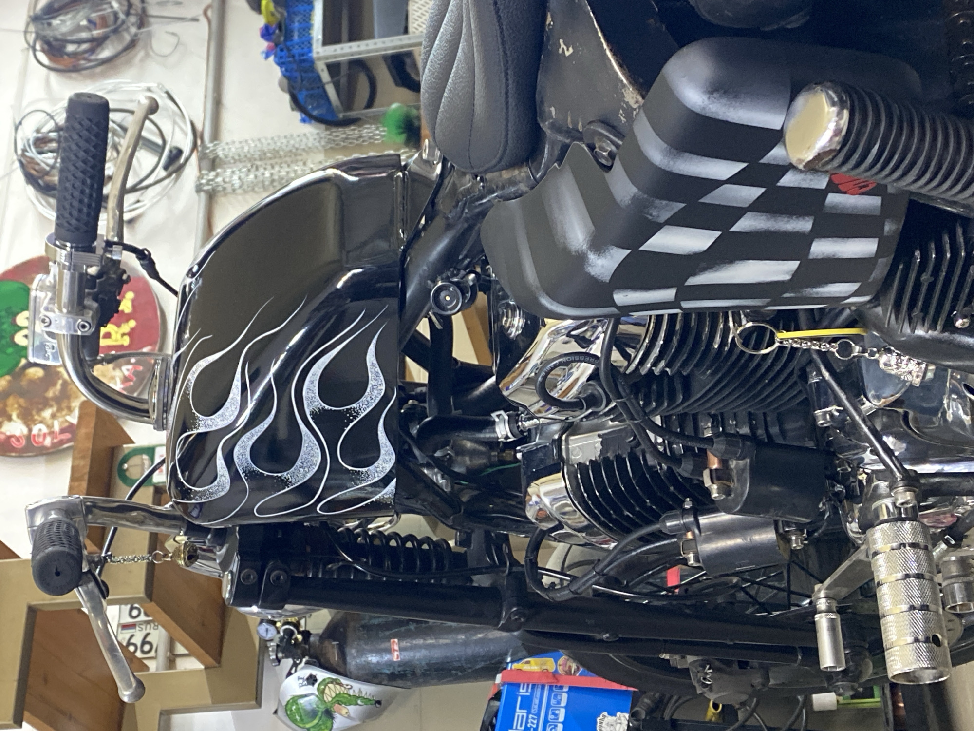 The image shows a close-up of a black motorcycle parked indoors, possibly in a garage or workshop. The motorcycle has decorative white flames on its fuel tank and a checkered pattern on a part of its body, likely a fender or side cover. Chrome accents are visible on various parts of the engine and controls. A small bell keychain hangs from the handlebars. The background includes shelves with items, electrical wiring, and a wall. The scene is brightly lit, suggesting artificial lighting. There is no indication of the city of Agronom, Russia, in the image.