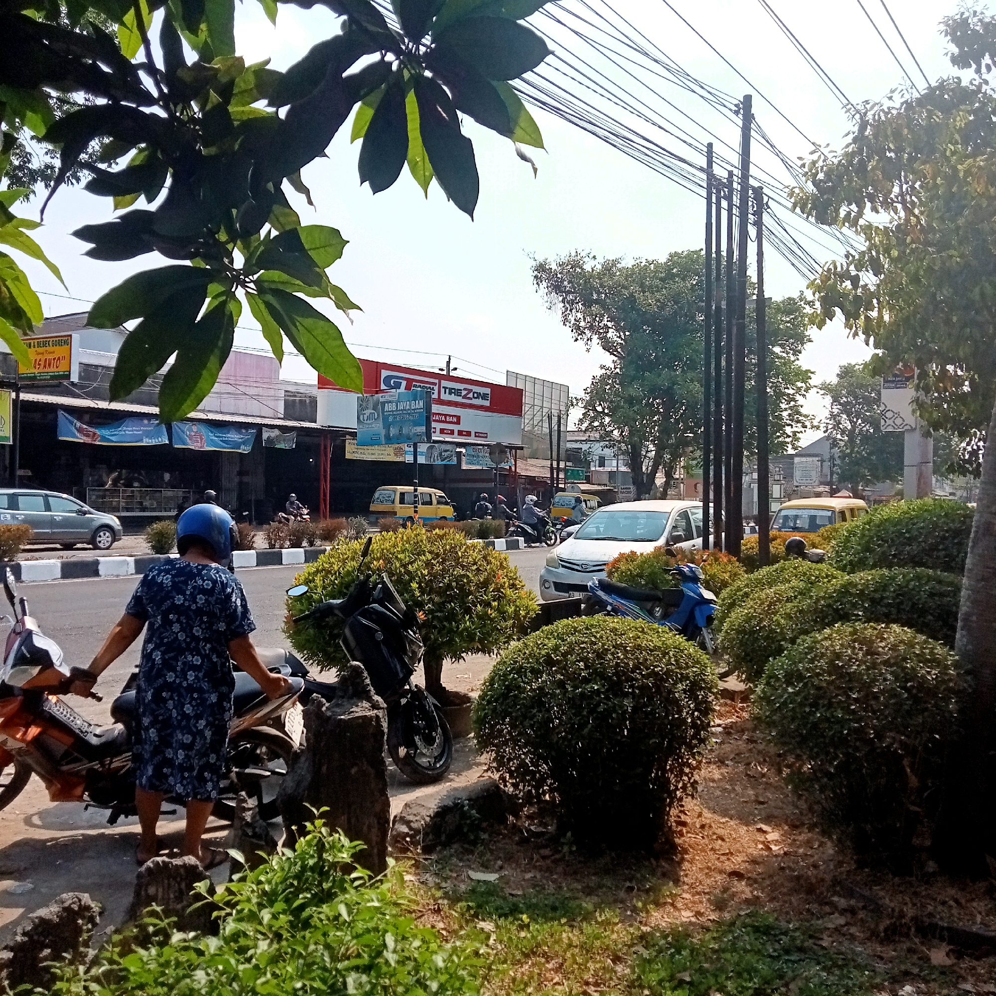 In Triyagan, Indonesia, on a sunny day, a person wearing a blue floral dress and a blue helmet is standing next to an orange motorcycle on a street. The person is facing away from the camera, holding onto the motorcycle's handlebars. To the left of the motorcycle, a grey sedan is parked on the side of the road. Across the street, a row of shops with banners are visible, including a "Tire Zone" establishment with a red and white sign. Several other vehicles, including yellow minivans and motorcycles, are present on the road and in the parking areas. Dense, well-maintained bushes are planted along the roadside, some in circular formations. In the background, electrical wires stretch across the sky, supported by poles. Trees with green foliage are scattered throughout the scene, and the sky is clear and bright.