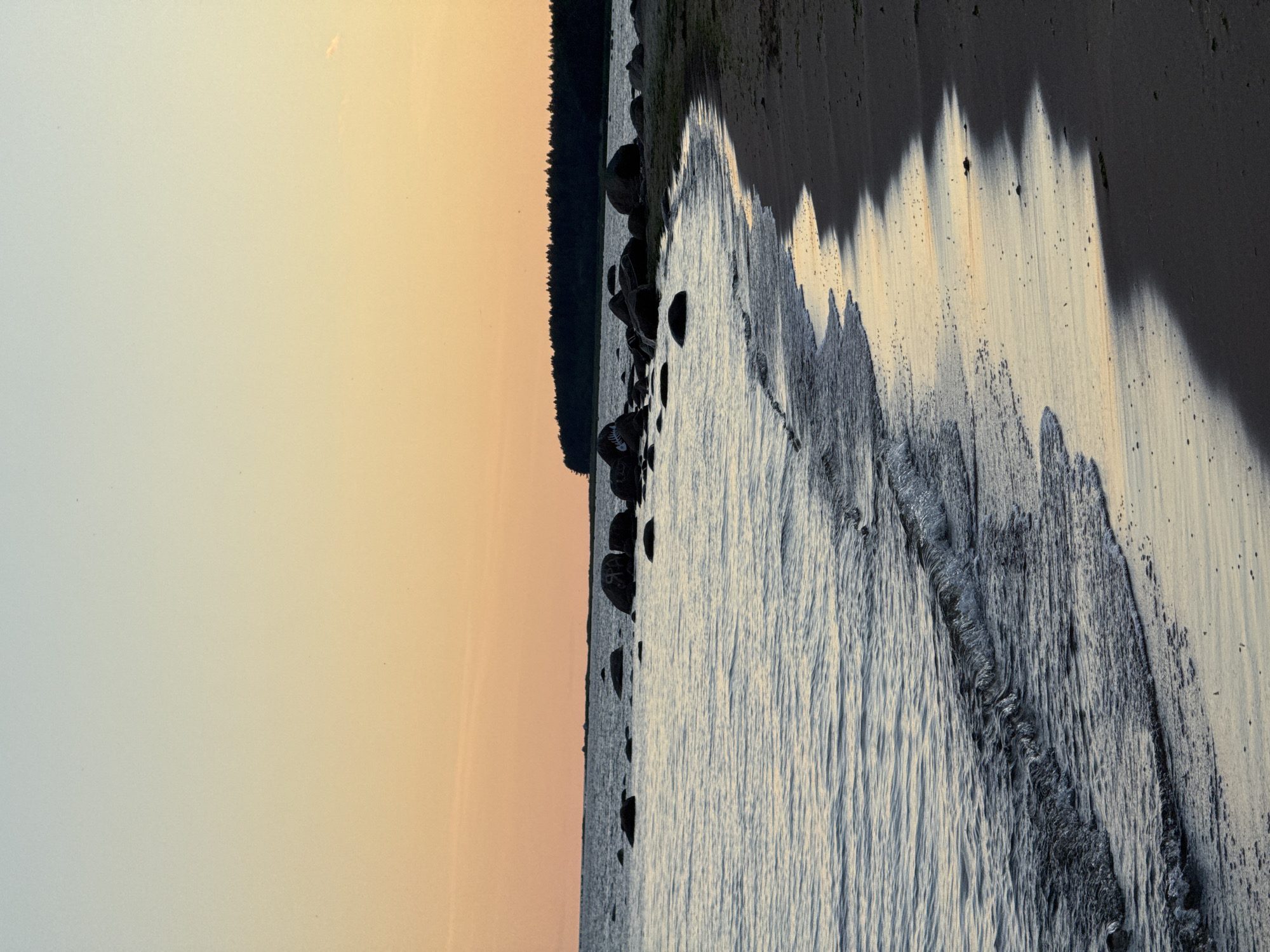The image shows a natural landscape at dusk. The sky transitions from a pale blue at the top to a warm orange hue near the horizon. A dark treeline forms the distant background, with a scattering of large, dark rocks visible in the water closer to shore. The water itself is calm, with small waves gently lapping at a sandy beach. The wet sand near the waterline reflects the colors of the sky, creating a band of warm tones against the darker, dry sand. There are no people or animals visible in the scene. The location is Zelenaya Roshcha, Russia.