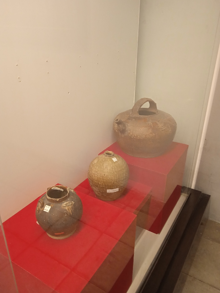 Three ceramic pots are on display behind glass in a museum in Surakarta, Indonesia, at the coordinates -7.558, 110.783. Two red pedestals support the pots. The leftmost pot is dark brown and has a small spout. The middle pot is light brown and appears to be textured. The rightmost pot is also light brown and has a large handle. All pots are displayed on red pedestals. The background is a white wall with small holes in it. The floor is made of dark brown wood. The glass case is clean and free of any visible dirt. The pots are well-lit and visible. The photo was taken from a slightly elevated angle.