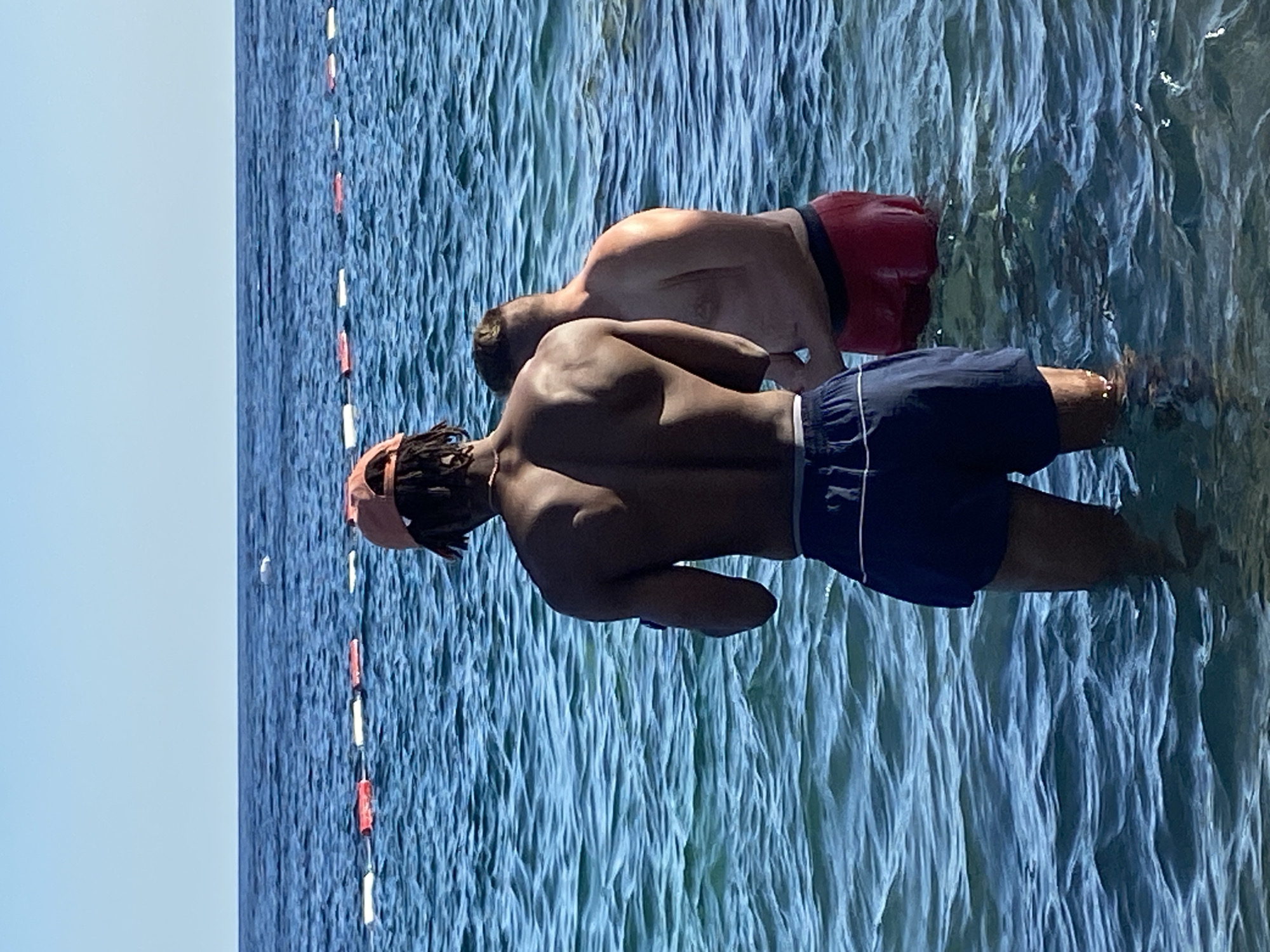 Two men stand in the clear blue water of the Adriatic Sea. The man in the foreground, facing away from the camera, has dark skin and wears a pink baseball cap and navy blue swim trunks. He has a lean, muscular build. To his right, also facing away, is a man with lighter skin, wearing red swim trunks. He has a fuller build. The water is calm with gentle ripples, and a line of red and white buoys stretches across the horizon in the distance. The sky above is a clear, bright blue. This scene is taking place in the waters off the coast of Prijevor, Montenegro, a small village near the popular tourist destination of Budva.