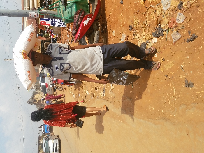 The image depicts an outdoor street scene in the city of Nnobi, Nigeria, under bright daylight. In the foreground, a male individual walks left-to-right, carrying a large white sack labeled "927-M" on his head and a black plastic bag in his left hand. He wears a grey short-sleeved polo shirt with dark blue pockets and collar, dark trousers, and dark flip-flops. To his left, slightly behind, a female individual walks away from the camera, wearing a red patterned top, a dark skirt, and light-colored flip-flops, carrying a small black handbag in her left hand.
The ground is unpaved, consisting of reddish-brown dirt with scattered debris, litter, and dark, irregular clumps. Shadows from the people and objects indicate strong overhead sunlight. In the mid-ground, a paved road is visible, with several vehicles, including a white mini-van and other cars, along with parked motorcycles. A blue vehicle is further down the road. Several other people are present in the background, some walking, some near vehicles. Utility poles with overhead power lines traverse the scene. On the right, a utility pole features a red and white sign with the letters "PU" prominently displayed, and a smaller sign partially visible below it. Various buildings, including a multi-story structure with a distinct roofline, are visible in the background, lining the street.