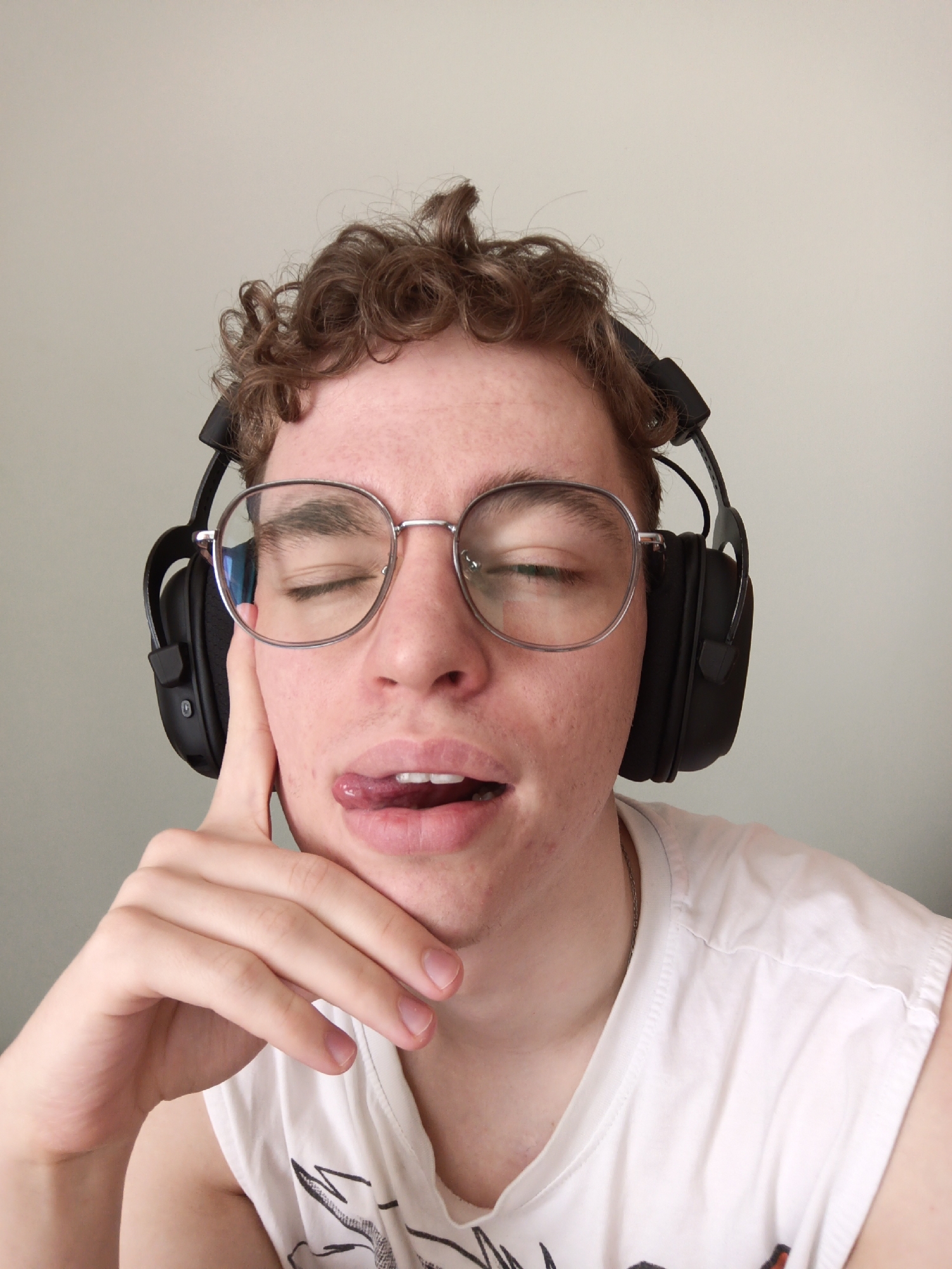A person with curly brown hair and wearing round, silver-rimmed glasses and black headphones is captured in a close-up shot. The person's eyes are closed, and their tongue is slightly extended from their mouth. Their hand is raised, with their index finger touching their cheek. They are wearing a white t-shirt. The background is a plain, light-colored wall. There is no visible evidence to indicate this photo was taken in Odintsovo, Russia, or any other specific location.