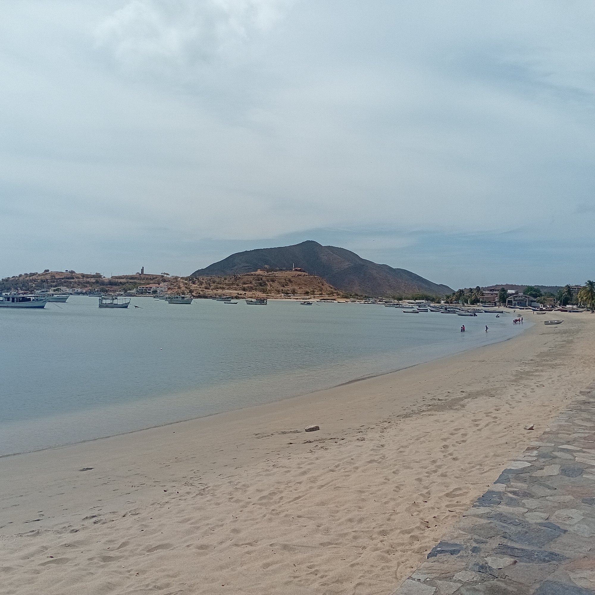 The image depicts a coastal scene in what appears to be Unknown, Unknown. The foreground features a sandy beach with footprints and a paved area of stone. The midground is dominated by a calm body of water with numerous small boats anchored or moored. A mountain rises in the center background, with buildings and structures visible on a lower hill to the left. A few people are seen on the beach and in the water towards the right side of the frame. The sky is overcast with light, gray clouds.