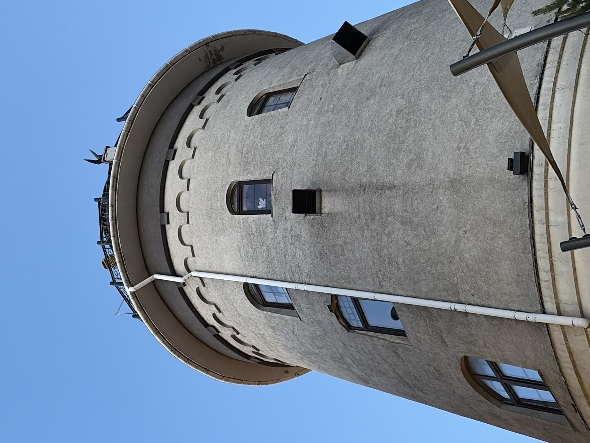 An upward-facing, wide-angle shot captures a portion of a cylindrical, grey, concrete tower against a clear blue sky. The tower has a textured, weathered surface and is adorned with several arched windows with dark frames. A white drainpipe runs vertically along the side of the tower. Near the top, decorative, scalloped stonework lines the base of a wide, circular cornice, which supports a dark metal railing. Black, decorative metal elements, resembling bird-like figures, protrude from the top edge. A beige awning is partially visible in the lower right corner, supported by a dark pole. The image is a low-angle perspective, emphasizing the towering structure.