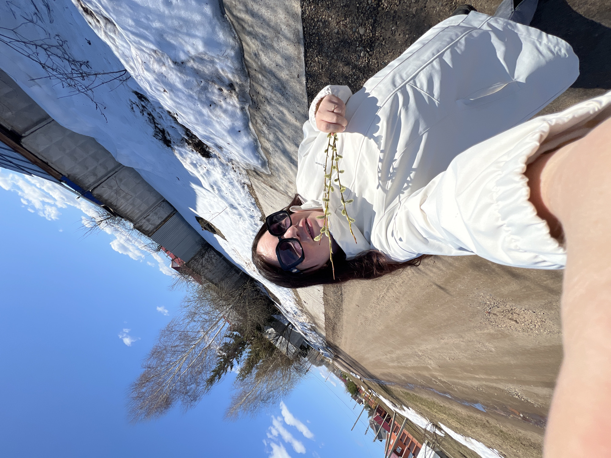A woman wearing a white coat and dark sunglasses is lying on a path with melting snow on either side, holding sprigs of willow. The scene suggests early spring in a residential area in Noviy, Russia, with a clear blue sky visible above buildings and trees.