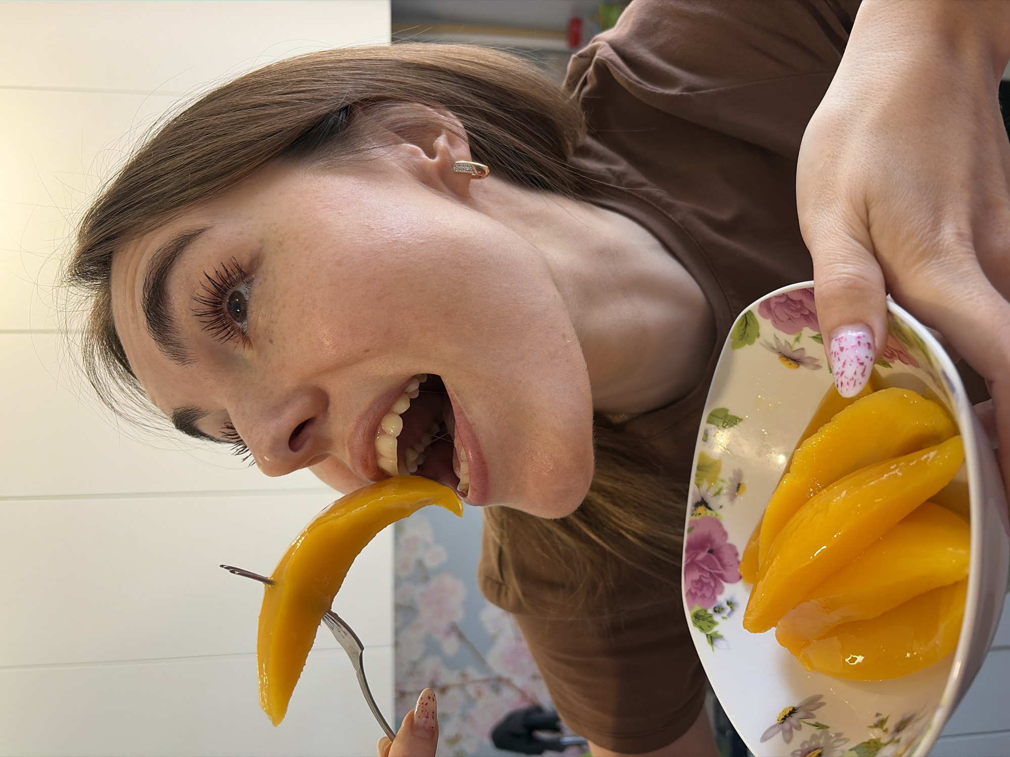 A woman with fair skin and brown hair is holding a plate of sliced mangoes and eating a piece off a fork. She wears a brown t-shirt and a gold earring. Her nails are manicured. The plate is white with a floral pattern. The background includes a white wall and wallpaper with a floral pattern. The location is in the city of Sarapul, Russia.