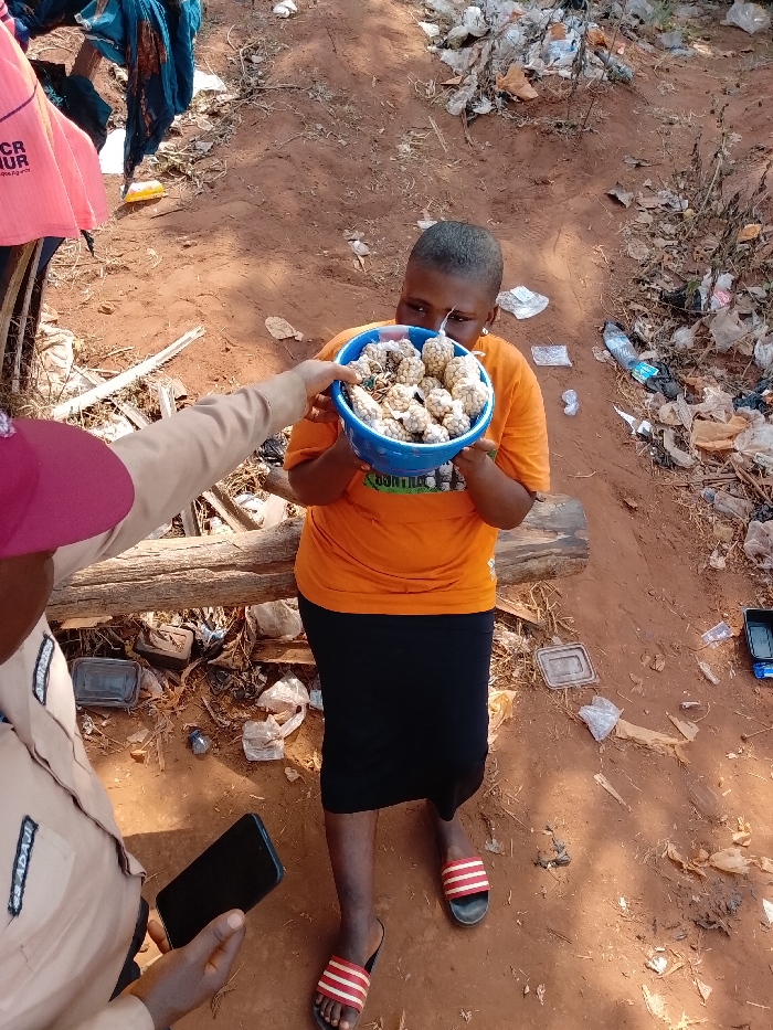 The scene depicts an outdoor interaction in Ejule, Nigeria, on unpaved reddish-brown dirt ground. A female subject, with short hair, wearing an orange t-shirt, a black skirt, and red-and-white striped slide sandals, stands centrally, holding a blue plastic bowl with both hands. The bowl contains numerous small, light-brown, roughly spherical items, many individually wrapped in small, light-colored mesh or net material. She is observed looking down into the bowl. From the lower-left foreground, a second individual, partially visible, wearing a light-colored uniform shirt with a "SS ADAVII" patch and a maroon cap, extends their right arm to present the bowl to the female. This individual concurrently holds a black smartphone in their left hand. The immediate environment is extensively littered with various discarded materials, including plastic bottles, plastic bags, and containers. Two horizontal wooden logs are visible behind the primary subjects, and colorful fabric hangs in the upper-left background.