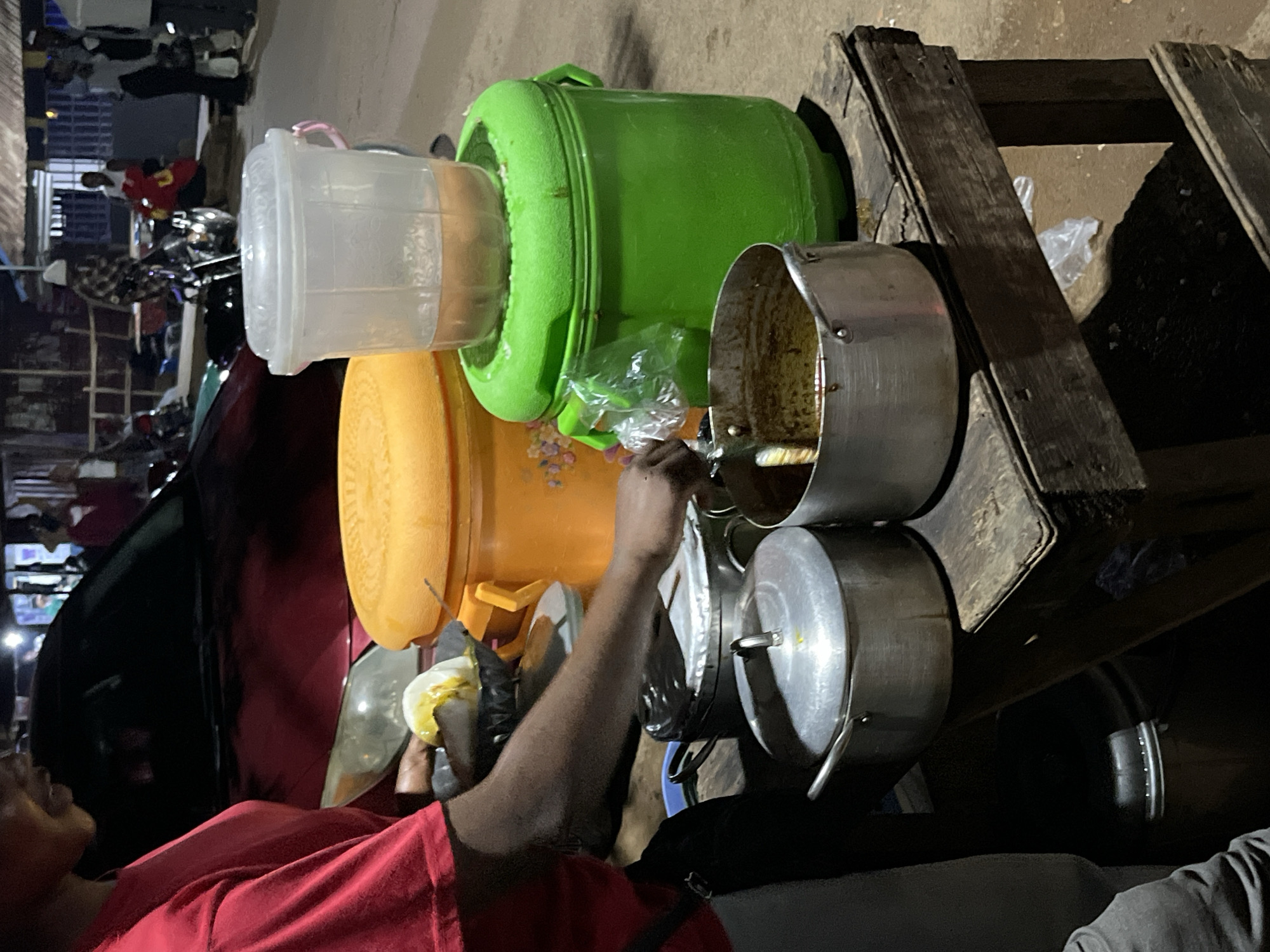 The image captures a bustling scene, likely a street food vendor in Barikin Gidar Kanat, Nigeria. The primary subject is a person wearing a red shirt, with their arm extended as they interact with a food preparation area. They appear to be holding a piece of food, possibly an egg, in a white bowl.
Surrounding the person are various containers and cooking implements. A large, bright green bucket is prominently featured, possibly used for storing ingredients or as a cooler. Next to it, an orange cylindrical container is visible, suggesting it holds more food items. Several metal pots are stacked, with one larger pot on the right appearing to contain some yellowish liquid, hinting at a dish being cooked or served. A transparent plastic container with a pink handle sits on top of the green bucket. The entire setup is placed on a rustic wooden structure, likely a makeshift cart or table, typical of street vendors.
The background shows a hint of a marketplace or a busy street with people and vehicles, including motorcycles, further reinforcing the setting. The lighting suggests it might be evening or night, given the artificial lights visible in the distance and the overall ambiance. The scene conveys a sense of everyday commerce and food preparation. There is no visible text in the image.