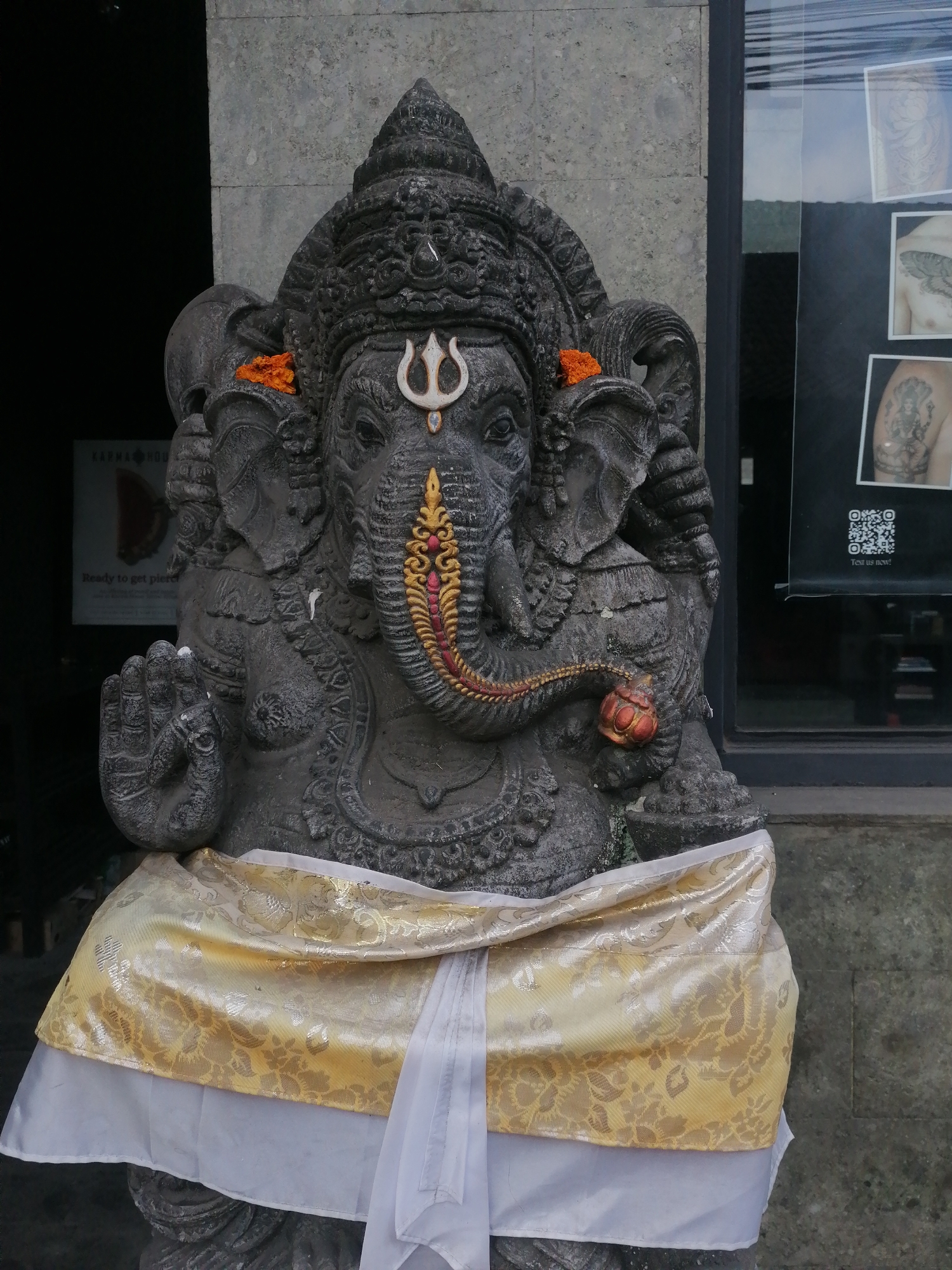 A dark stone statue of Ganesha, a Hindu deity with an elephant head, stands in front of a building. The statue is adorned with an orange flower on its head and a yellow and white patterned cloth around its waist. Behind the statue, a stone wall is visible, and to the right, a glass window displays several framed images of tattoos. A QR code is also present on the window. A sign with text is visible to the left of the statue. The scene appears to be outdoors, possibly on a street in Ubud District, Indonesia.