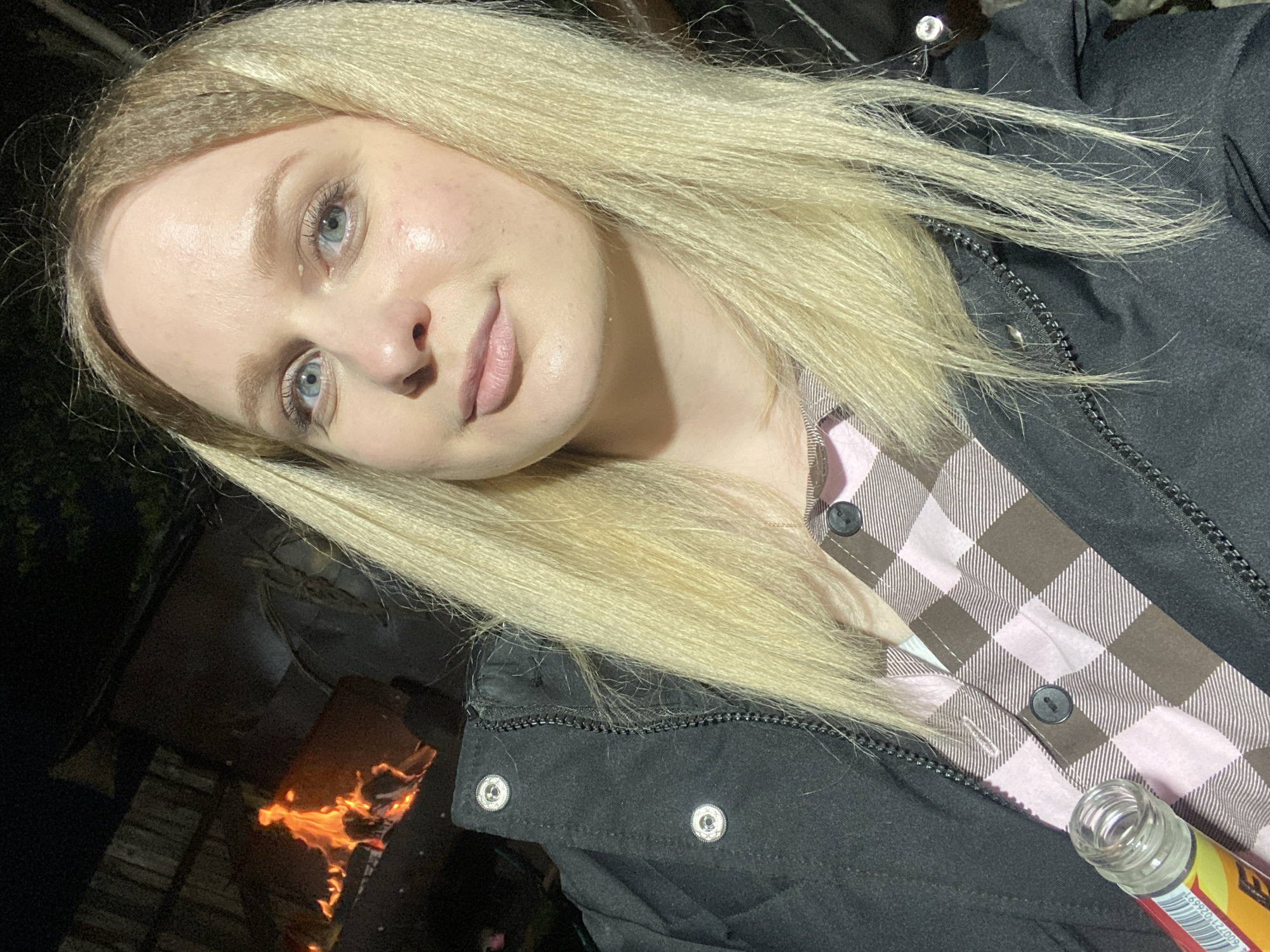 A close-up selfie, captured at night in Kauchuk Stantsiya, Russia, features a young woman with fair skin, blonde hair, and light blue eyes. She is seen from the chest up, tilting her head slightly, and looks directly at the camera with a subtle, neutral expression. She is wearing a black jacket with visible snaps and a zipper, layered over a pink and brown plaid button-up shirt. Her blonde hair frames her face and extends over her shoulders and jacket.
In her right hand, a clear glass bottle with a yellow and red label is visible, partially obscured, showing the text "169920" and parts of "EN" on its label. The background is dark, indicating nighttime, with a bright, active fire, featuring glowing embers and flames, visible in the lower left, suggesting an outdoor setting or bonfire. The warm clothing worn by the woman implies cool or cold weather.