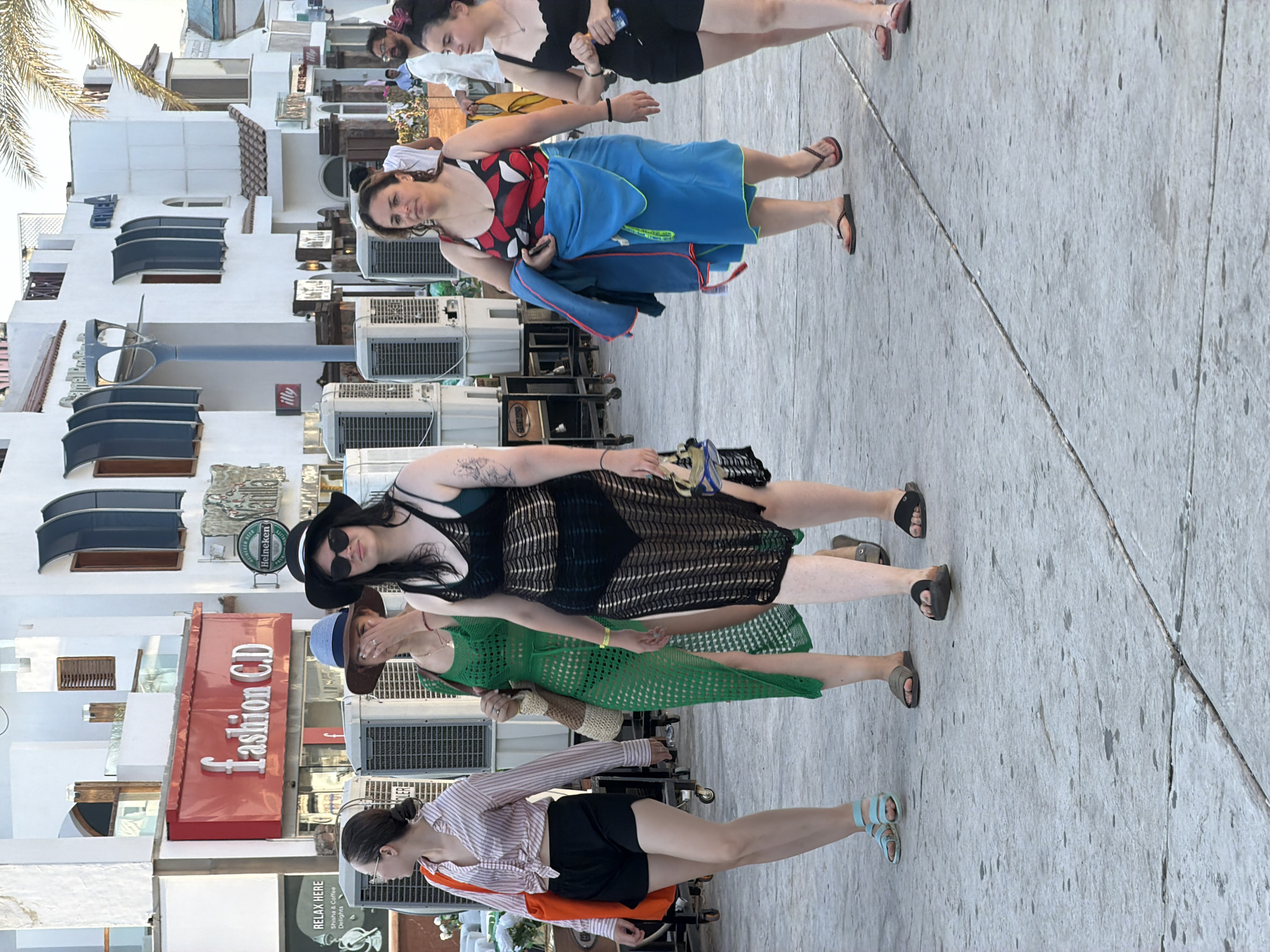 A group of five women are walking along a paved promenade in Sharm El Sheikh, Egypt. The buildings in the background are white with blue awnings and some signage, including "fashion C.D." and "illy." The women are dressed in summer attire, including swimsuits, cover-ups, shorts, and sandals. The woman in the foreground on the left is wearing a pink and white striped shirt, black shorts, and blue sandals. Next to her, a woman in a green crochet dress and brown sandals is walking. Another woman in a black mesh cover-up over a swimsuit and black sandals is in the center. To her right, a woman in a red, white, and black swimsuit and a blue towel wrapped around her waist is walking. The woman on the far right is wearing a black tank top, black shorts, and black sandals. There are large air conditioning units lined up along the buildings. The lighting suggests it is daytime, possibly late afternoon.