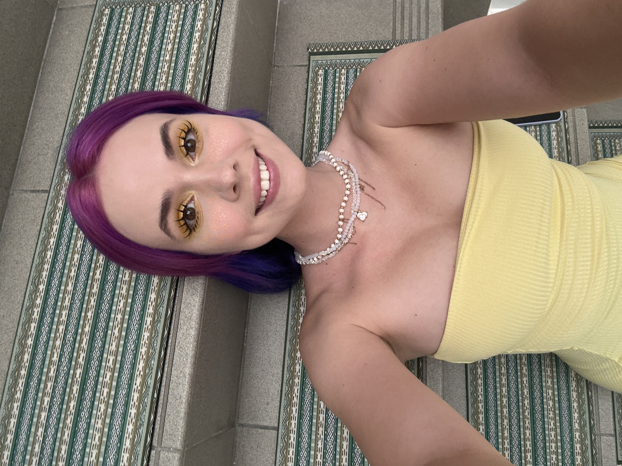 A woman with purple hair and yellow makeup lies on a patterned mat on a gray concrete staircase. She wears a yellow strapless dress and a layered beaded necklace. The location is Zagreb, Croatia.