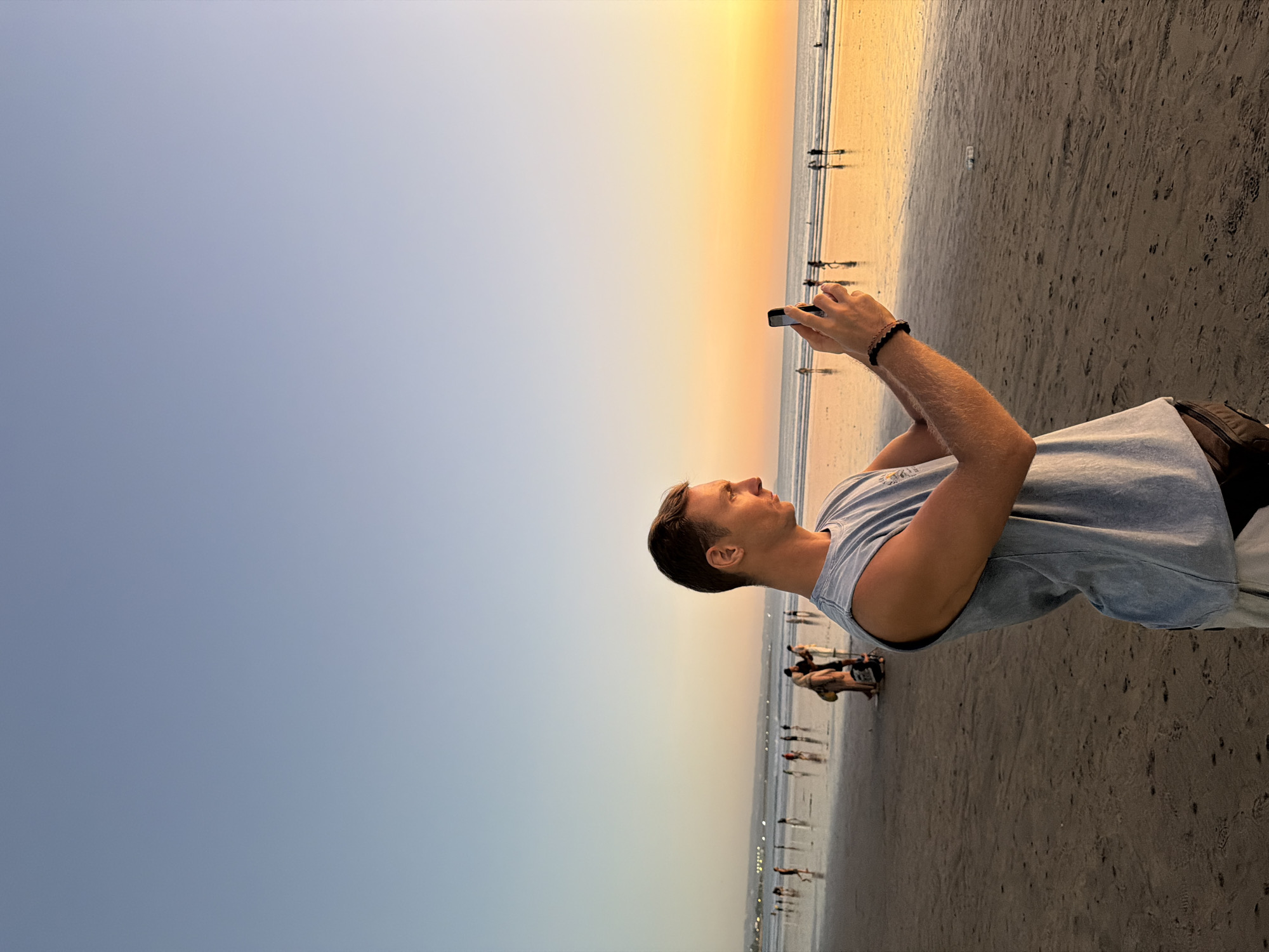 A man stands on the wet sand of Kuta Beach, Indonesia, holding a smartphone horizontally to take a photograph. He is wearing a light blue sleeveless t-shirt and dark shorts. His left arm is extended, holding the phone, and his right hand is on the side of the phone, also holding it. He is looking towards the phone, his profile visible. In the background, the ocean stretches to the horizon, with a warm sunset casting an orange glow across the sky and reflecting on the wet sand and water. Several other people are scattered across the beach, some walking, others standing, their figures silhouetted against the vibrant sunset. The sky transitions from a pale blue at the top to a rich orange near the horizon.