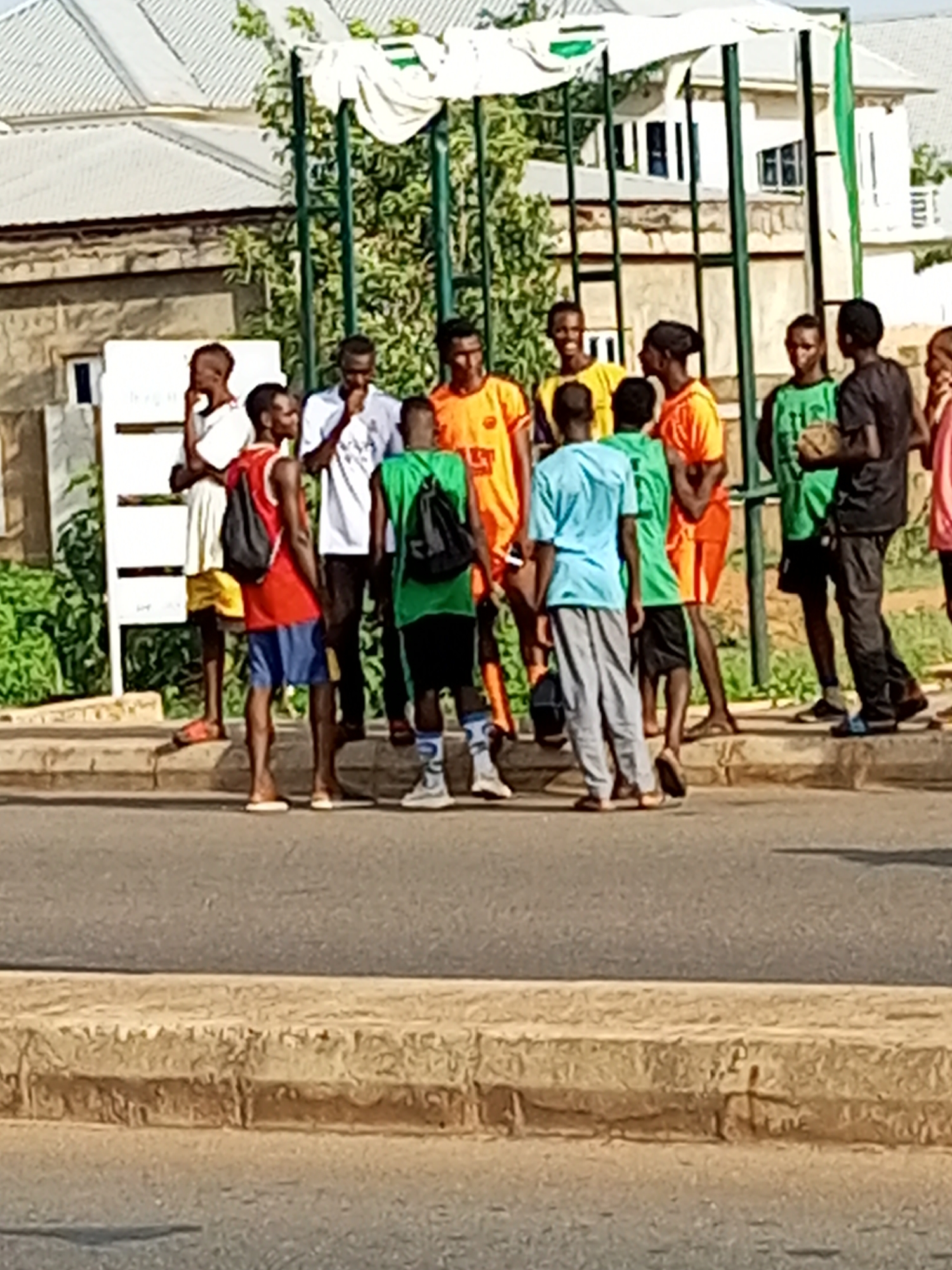In Gombe, Nigeria, a group of young men stand on a paved road, with a stone curb in the foreground. They appear to be near a sports field or park, indicated by a green net-like structure and some trees. Most of the men are dressed in athletic wear, with several wearing brightly colored jerseys and shorts. One man in a red tank top and blue shorts carries a black backpack. Another man in a green shirt holds a small, round object, possibly a ball. The men are standing and interacting, some looking at each other while others gaze in different directions. Behind them, buildings with distinctive white roofs and textured walls are visible, along with a white signpost. The overall scene suggests a casual gathering, possibly before or after a sporting activity.
