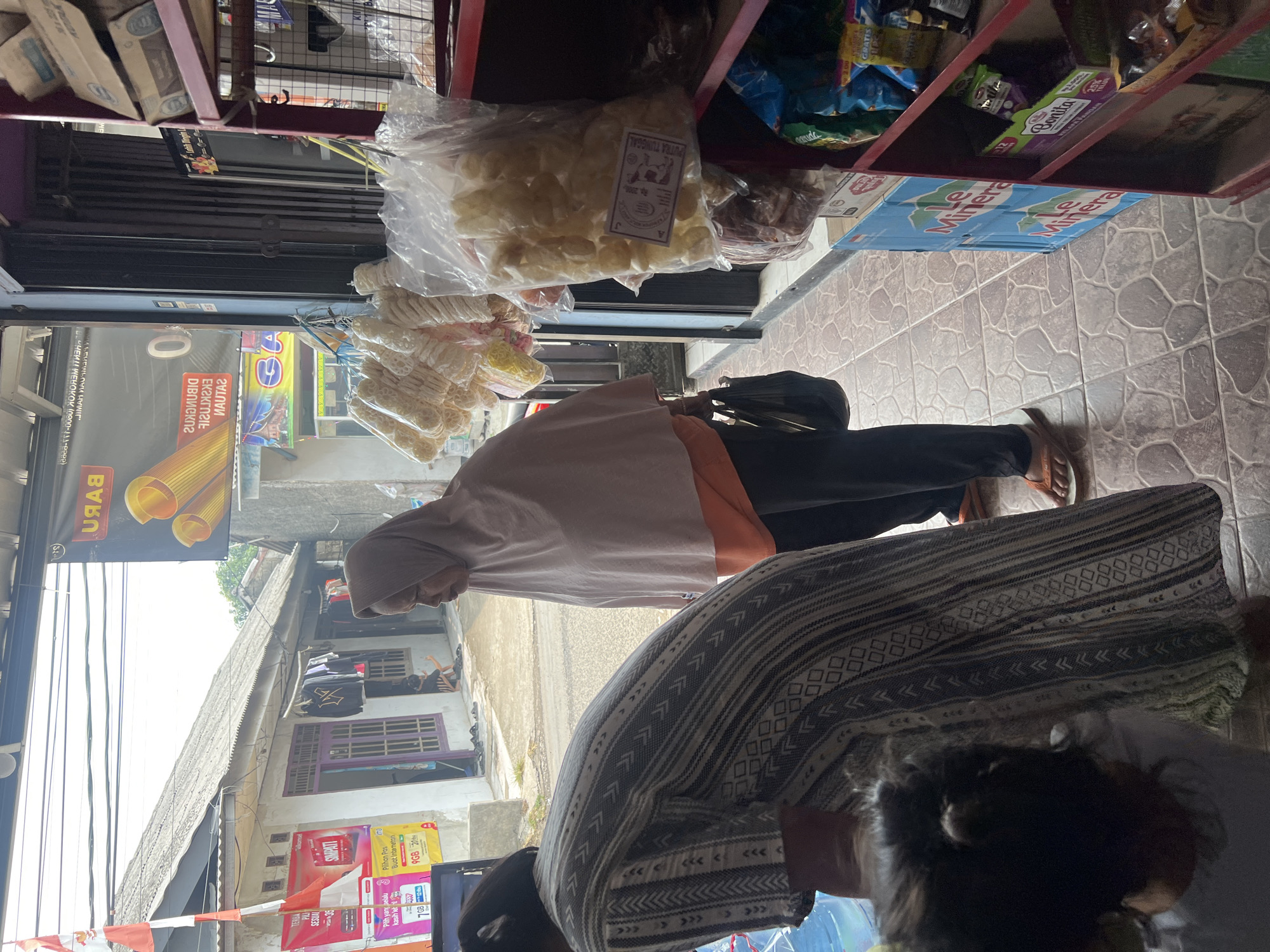 The image depicts a street-level view of a small retail establishment in Klapanunggal, Indonesia. A woman wearing a light pink hijab and an orange shirt is standing inside the doorway of the shop, facing right. She is holding a black bag in her left hand. In the foreground, another person, partially obscured and facing away from the camera, is wearing a patterned dress. This individual appears to be interacting with items on a table or counter. Behind the woman in the hijab, the interior of the shop is visible, with shelves stocked with various goods, including bagged snacks. The exterior shows a residential building with a tiled roof and posters on the wall. The ground is paved with patterned tiles. The scene suggests a typical commercial transaction in a local Indonesian community.