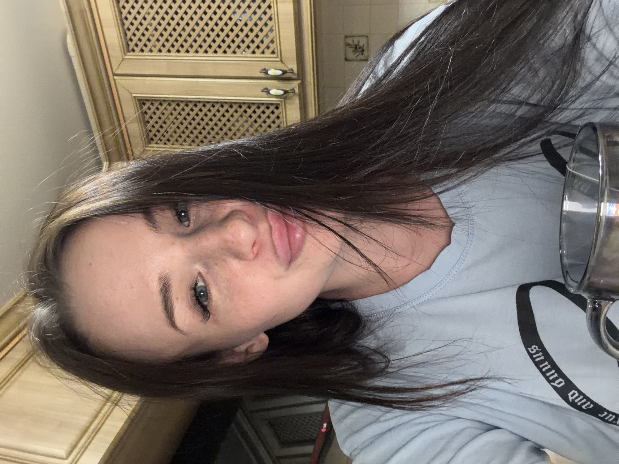 A young woman is prominently featured in this close-up, rotated image, likely a selfie, taken indoors. Her head is tilted, and she looks directly towards the camera with a neutral to slightly smiling expression, her lips slightly parted. She has long, dark brown hair, with some lighter strands visible around her face and on top of her head. Her skin shows a scattering of freckles. Her eyes are blue. She is wearing a light blue, long-sleeved top.
In the background, behind her, are wooden cabinets with intricate latticework panels and decorative, possibly ceramic, handles. The cabinets appear to be light-colored wood. To the right of the cabinets, a small decorative tile is affixed to a tiled wall. Part of a metallic, possibly chrome, mug or cup with a handle is visible in the lower right corner, obscuring some of her shirt.
On the black stripe of her light blue shirt, there is visible white text. Due to the image's rotation, the text appears upside down and reads: "snnuG dnA sunnO".
The setting appears to be an indoor space, possibly a kitchen or dining area, given the style of the cabinetry. The lighting suggests artificial indoor illumination. The time of day and weather are not discernible.