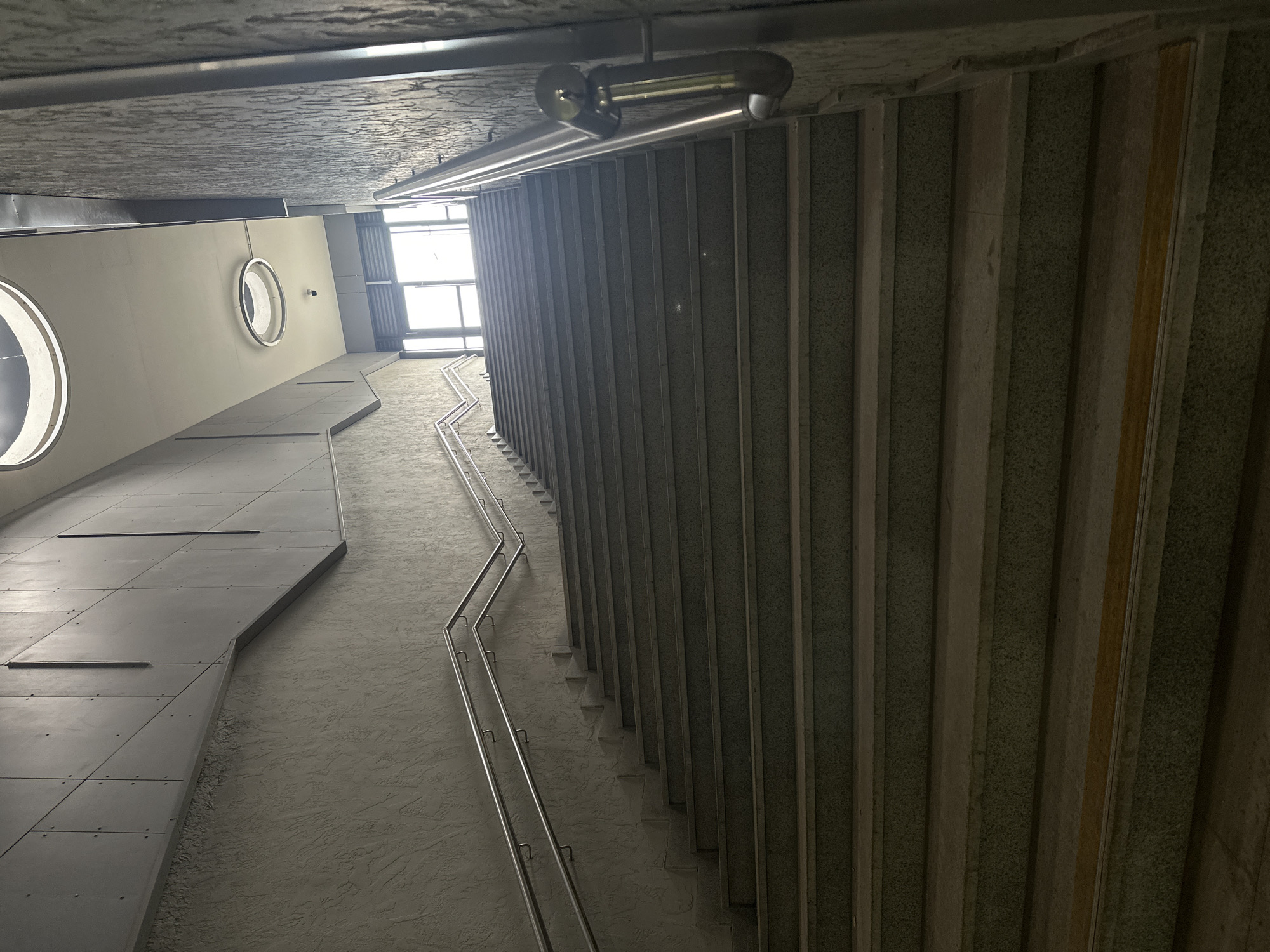 This interior shot depicts a modern architectural space with contrasting elements. On the left, a smooth, flat, metal-clad wall features circular windows. Parallel to this, on the right, a textured, vertical wall composed of alternating panels creates a sense of depth. A metal handrail snakes down a set of stairs between the two walls, leading towards a bright window in the distance. Above, a concrete ceiling is visible, and a silver cylindrical light fixture is mounted to it.
This image appears to be of the interior of the Kazan Metro's Kremlyovskaya station located in Kazan, Russia.