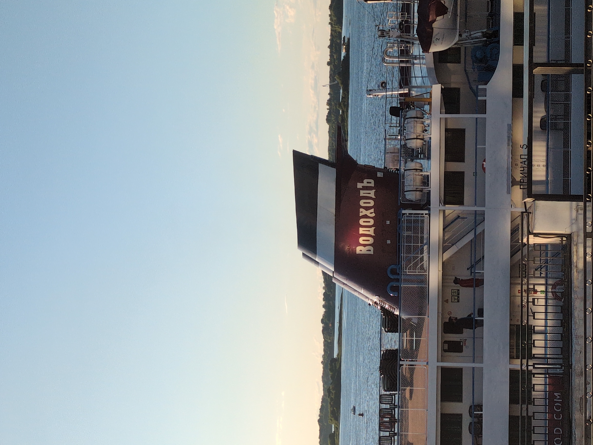 The image shows a close-up view of a ship docked on a body of water, likely a river, with a clear sky and a treelined shore in the background. The ship appears to be a passenger vessel, with multiple decks and visible lifeboats. The setting suggests a calm day, possibly late afternoon or early evening due to the warm, soft light.
The most prominent feature is the stern of the ship, which is painted a deep maroon color. White Cyrillic lettering on the stern spells out "ВОДОХОДЬ", which translates to "Water-Walker" or a similar term for a river vessel. To the right of the stern, the side of the ship is visible, showing windows, railings, and more white lettering that reads "ПРИЧАЛ 5", indicating "Berth 5".
Inside the ship's structure, through the windows, several people can be seen. One person is wearing a red jacket, and another appears to be a man with dark hair. The presence of people suggests the ship is either preparing for or has recently concluded a journey, or perhaps is docked for a period. The overall atmosphere is one of tranquility and possibly anticipation. The presence of the ship and the Cyrillic text strongly suggest a location in Russia, specifically hinted at by "Nizhny Novgorod" in the location context.