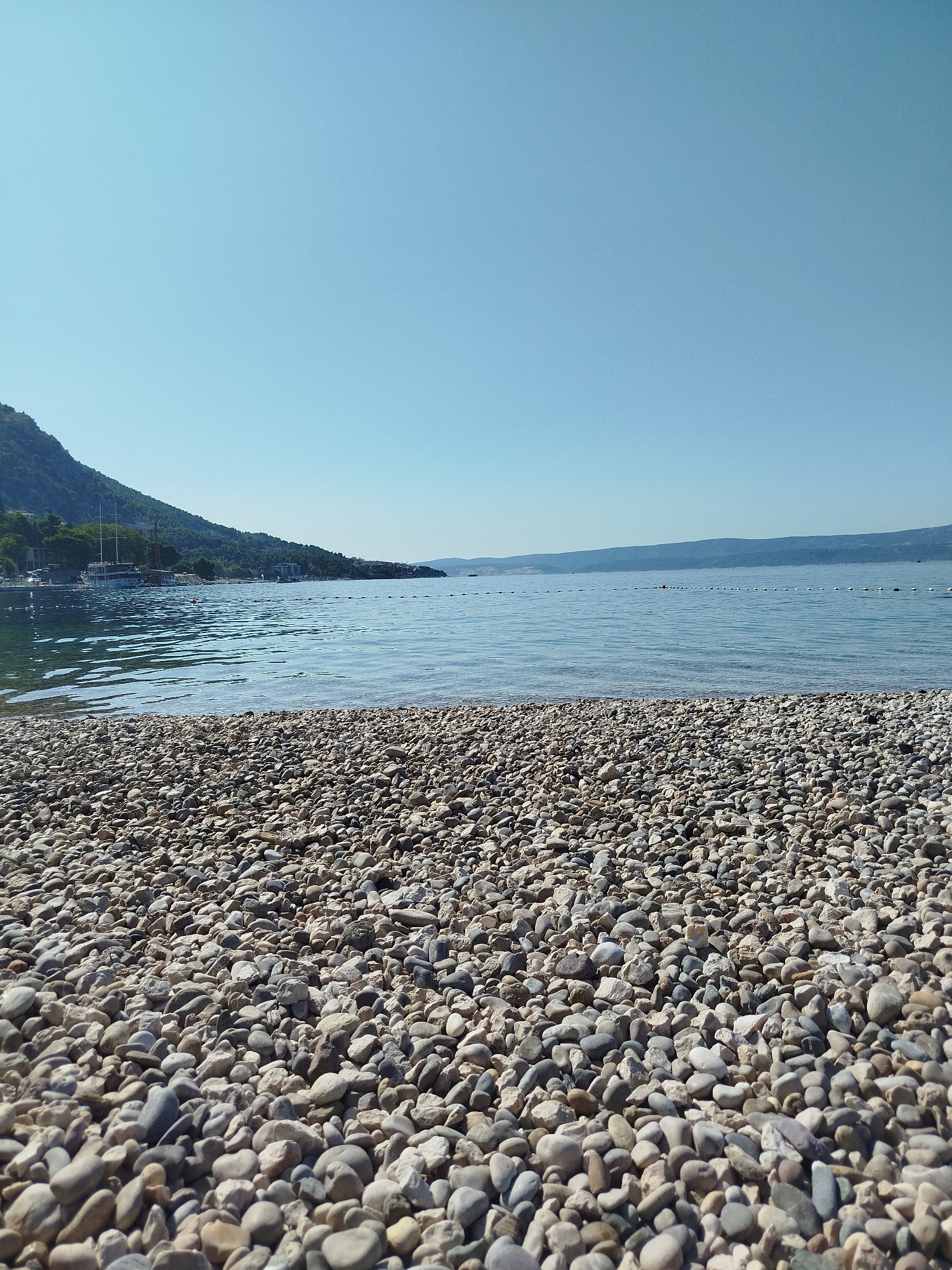 A wide-angle, low-angle view of a pebble beach in Omiš, Croatia, on a clear, sunny day. The foreground is filled with smooth, multicolored pebbles of varying sizes. The sea stretches out beyond the beach, its surface gently rippling under the sunlight. In the mid-ground, a line of buoys floats on the water, marking a boundary. Along the distant shoreline, lush green hills rise, dotted with buildings and trees. The sky above is a clear, pale blue, devoid of clouds. There are no people or animals visible in the scene.