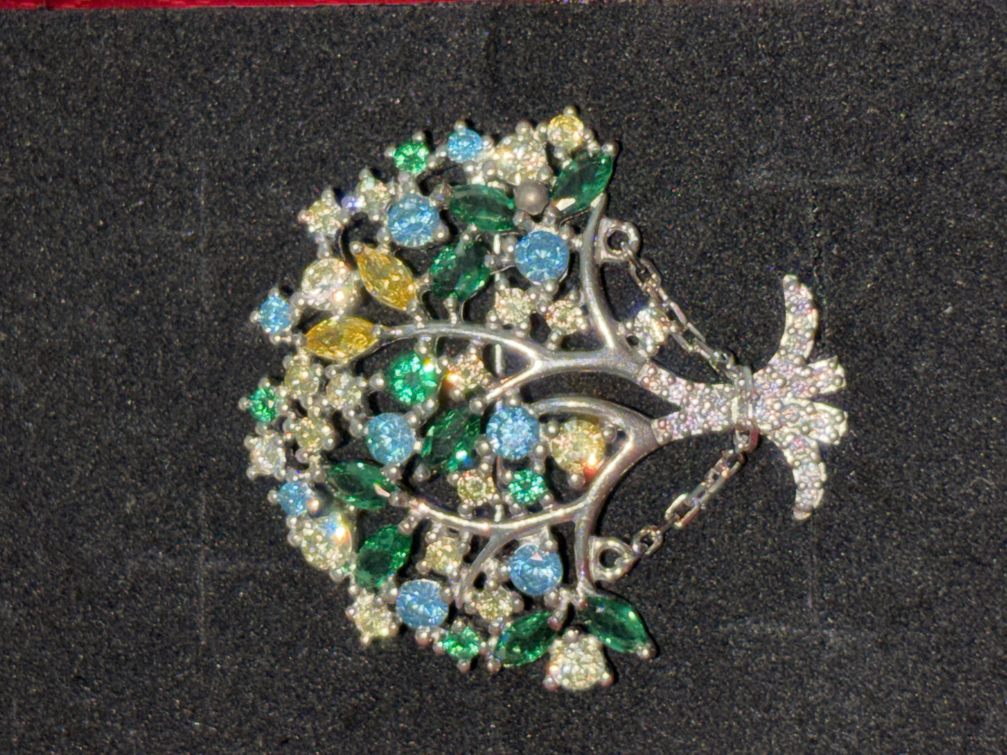 A close-up shot shows a silver brooch designed as a tree with colorful gemstones. The brooch is set against a dark, textured background. The tree has a silver trunk and branches adorned with various green, blue, and yellow gemstones, resembling leaves and fruit. Some of the gemstones are round, while others are marquise-shaped. The brooch has a small silver chain attached to the bottom. The lighting highlights the sparkle of the gemstones and the metallic sheen of the silver. The overall impression is of an intricate and decorative piece of jewelry.