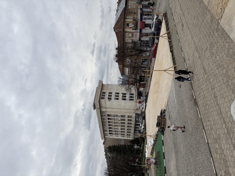 An elevated view captures a public square in Cherven Bryag, Bulgaria, on an overcast day. A large, rectangular, ground-level structure with a light-colored, possibly gravel or sand, surface occupies the midground. This structure is bordered by wooden framing and supported by several wooden tripod braces, indicating ongoing construction or temporary installation. To its immediate left, a smaller area features green turf, stacked wooden pallets, white material bags, and a display sign.
The foreground consists of a paved surface with hexagonal tiles and cobblestones. Three pedestrians are visible: one individual walks left-to-right across the cobblestones wearing light-colored pants and a light jacket, while two others walk right-to-left closer to the temporary structure, dressed in dark clothing.
In the background, a multi-story, light-colored building, identified by a banner as "ОБЩИНА ЧЕРВЕН БРЯГ" (Municipality of Cherven Bryag), stands on the left, with several cars parked in front. To the right, a row of older buildings with tiled roofs lines a street, where additional cars are parked. One building clearly displays "Пощенска Банка" (Postbank) signage and a small Bulgarian flag. Bare deciduous trees are scattered throughout the scene.