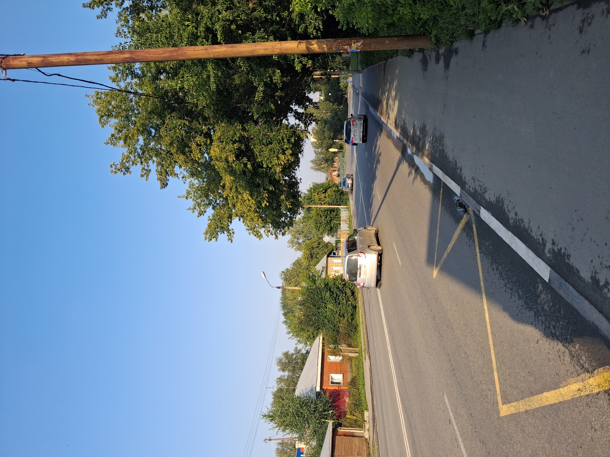 The image captures a sunny day on a street in Novosibirsk, Russia, likely during the late afternoon given the long shadows. The scene is dominated by a two-lane asphalt road with double white lines indicating traffic lanes. Yellow lines create a bus stop zone on the right side of the road, marked by a diamond shape and a solid yellow line. A light-colored minivan is driving away from the camera in the right lane, while another car is visible further down the road.
The right side of the road features a gray concrete sidewalk with a row of green bushes lining its edge. A tall, rusty utility pole stands prominently on the sidewalk, with wires running from it. To the left of the road, several houses with wooden siding and pitched roofs are visible, interspersed with lush green trees and smaller bushes. A few more cars are parked or driving on the road in the distance. The sky is a clear, bright blue. The overall impression is of a typical suburban street with residential buildings and active traffic under a clear sky.