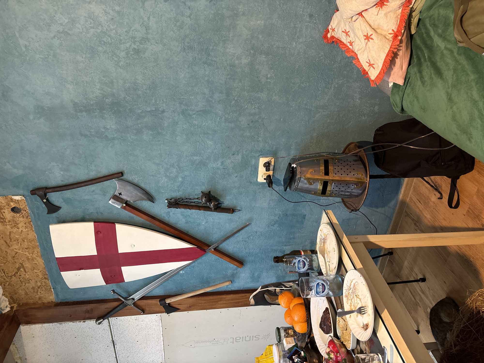 This image depicts an indoor space, likely a home in Verinsko, Bulgaria, featuring a blend of rustic and medieval-inspired decor alongside a functional dining area. A textured, teal-blue wall dominates the left and center, serving as a display for a collection of faux medieval weapons. These include a white shield with a red St. George's Cross, several axes of varying designs (one with a small cross symbol), a long sword, and a flail with a spiked ball.
Below the wall art, a light-colored wooden table is set, suggesting a recent meal. It holds multiple plates with food remnants, forks, two clear drinking glasses, and bottles. The glasses and a plastic bottle prominently feature the Cyrillic text "БАНКА" (Bank), a common mineral water brand in Bulgaria, confirming the location context. A bowl of oranges and other jars and bowls containing food items are also on the table.
In the lower right, a brown and white cat is partially visible, resting on the floor near some straw or hay, under the table. An electrical outlet is mounted on the blue wall, next to which a metal object resembling a medieval helmet or chest piece rests on a stand, with wires connected. A black backpack is also visible nearby.
In the upper right corner, a light-colored blanket with orange dragonfly patterns and fringes, along with a green blanket, are draped over an unseen piece of furniture, possibly a bed or chair. The lower left of the image reveals a section of unfinished drywall with the partially visible text "Rigips" and "Professional", indicating either ongoing construction or a deliberate aesthetic. The scene conveys a lived-in, informal atmosphere.
