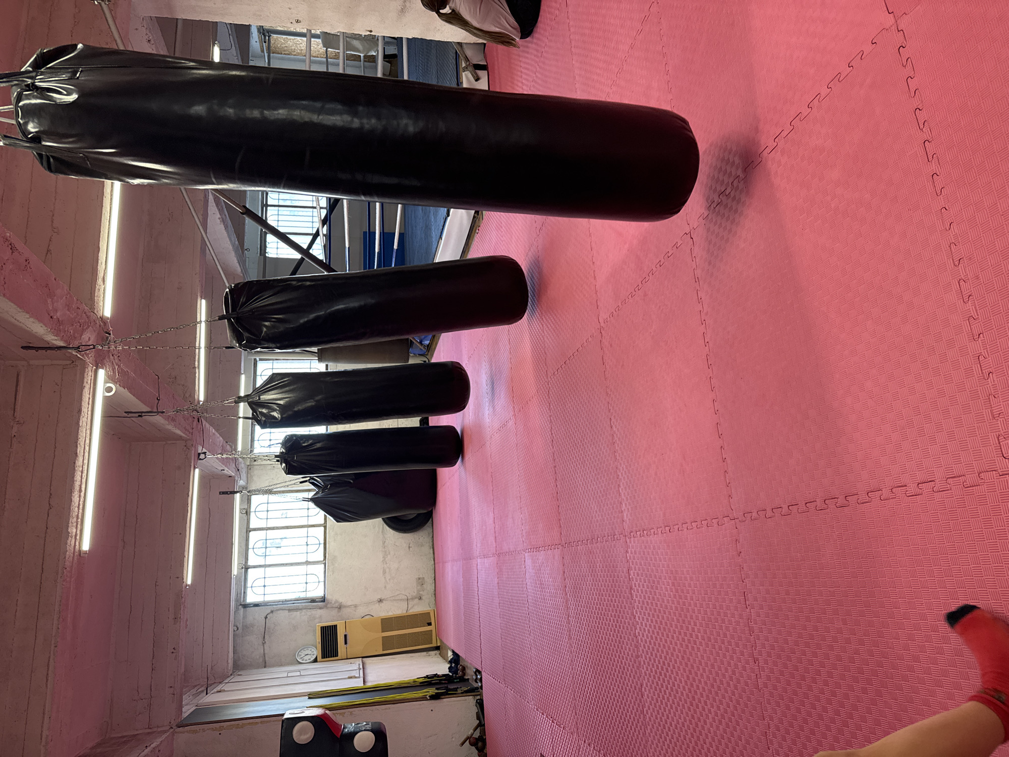 The image shows a boxing gym in Varna, Bulgaria. The floor is covered in pink interlocking foam mats. Several black punching bags of varying sizes are suspended from the ceiling by chains. There is a person in the upper right corner with a red sock. A window is visible on the left side of the frame, along with a heater. Other gym equipment is visible in the lower left corner.