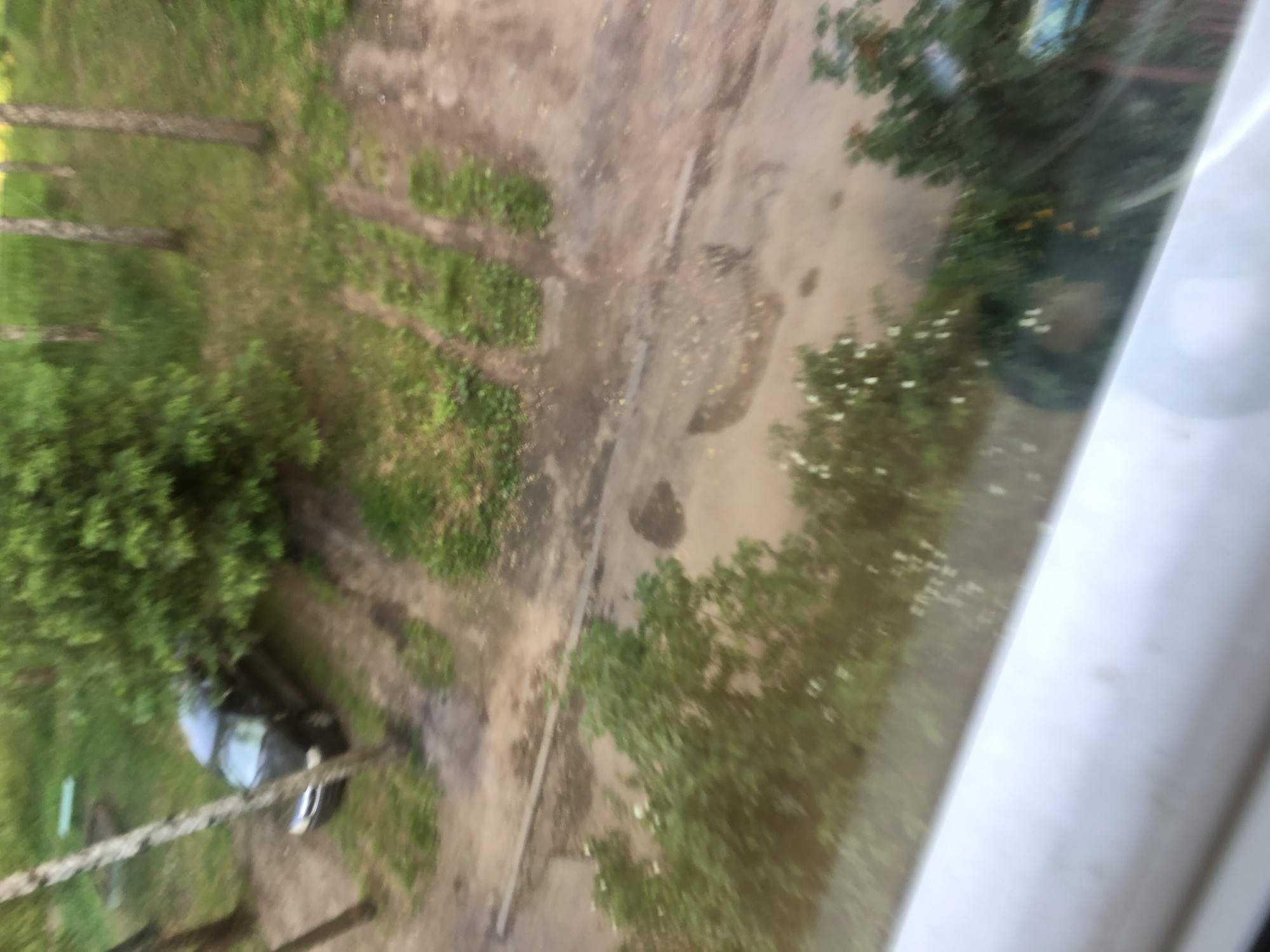 The image, taken from an elevated perspective possibly a window, shows a dark-colored vehicle parked on a dirt path adjacent to a grassy area. Trees are visible surrounding the car and in the background, suggesting an outdoor, possibly residential or park-like setting within the city of Safonovo, Russia. The ground appears to be a mixture of dirt, grass, and some puddles or wet patches, indicating recent rain or damp conditions. The overall impression is of an overcast day, with the lighting appearing diffused. The car's make and model are not clearly discernible due to the image's blurriness and distance. There are no people visible in the frame.