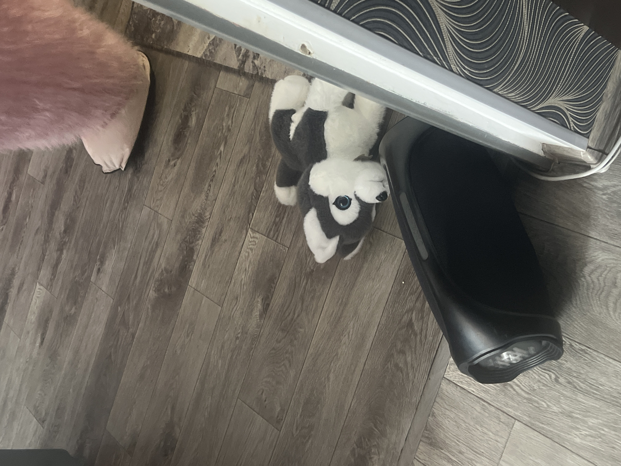 An indoor scene is depicted, featuring a light brown laminate wood floor with visible grain patterns. In the upper right quadrant, a light pink, densely furred leg terminates in a smooth, light pink slipper, indicating the presence of a foot. A vertical, light grey structural element, consistent with a door frame or jamb, borders the right side of the image. Adjacent to this structure, a wall is covered with dark wallpaper featuring intricate, swirling light-colored patterns. A white electrical cable runs along the base of the structural element near the bottom right corner. On the floor, towards the center-left, a black and white husky-dog plush toy with light blue eyes rests on its back. Below the plush toy and closer to the foreground, a black, cylindrical portable speaker lies horizontally on the floor, its visible end featuring a recessed circular grille. No specific actions or interactions are observed between the subjects. The location is an unspecified interior space in Unknown, Unknown.