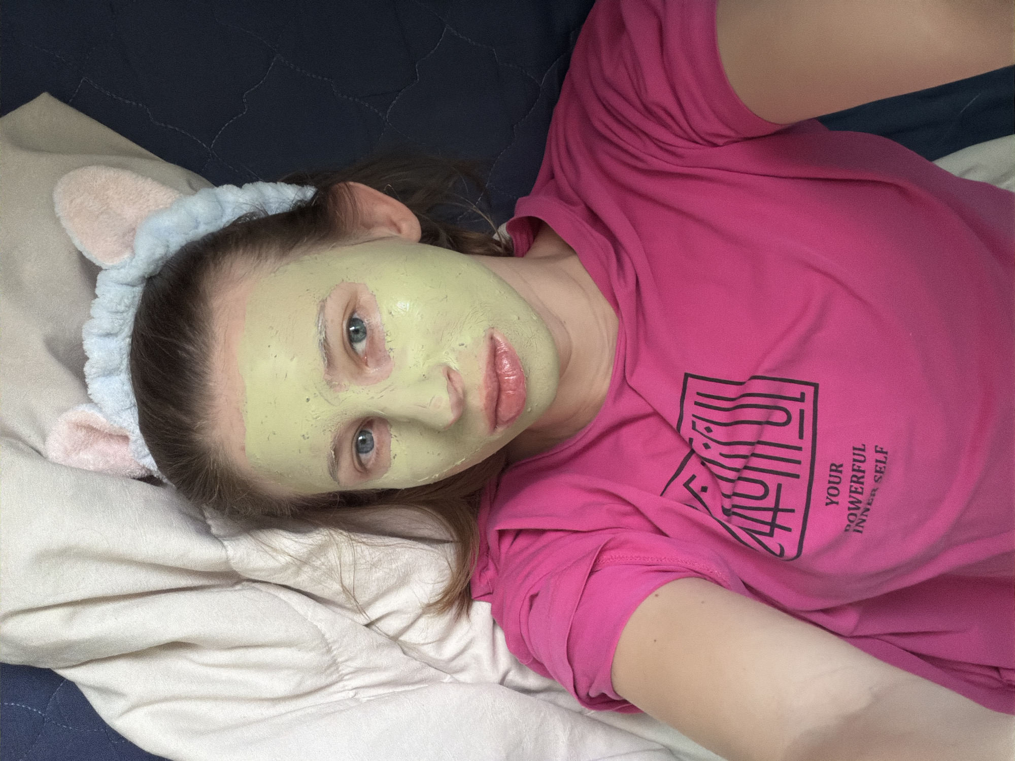 A young woman with a green face mask and a pink and blue fuzzy headband with cat ears is lying down on a bed. She is wearing a bright pink t-shirt with black text that reads "YOUR POWERFUL INNER SELF". The environment appears to be a bedroom in Dongducheon-si, South Korea, with a white duvet and a dark blue bedspread visible. The lighting is natural, suggesting it may be daytime.
