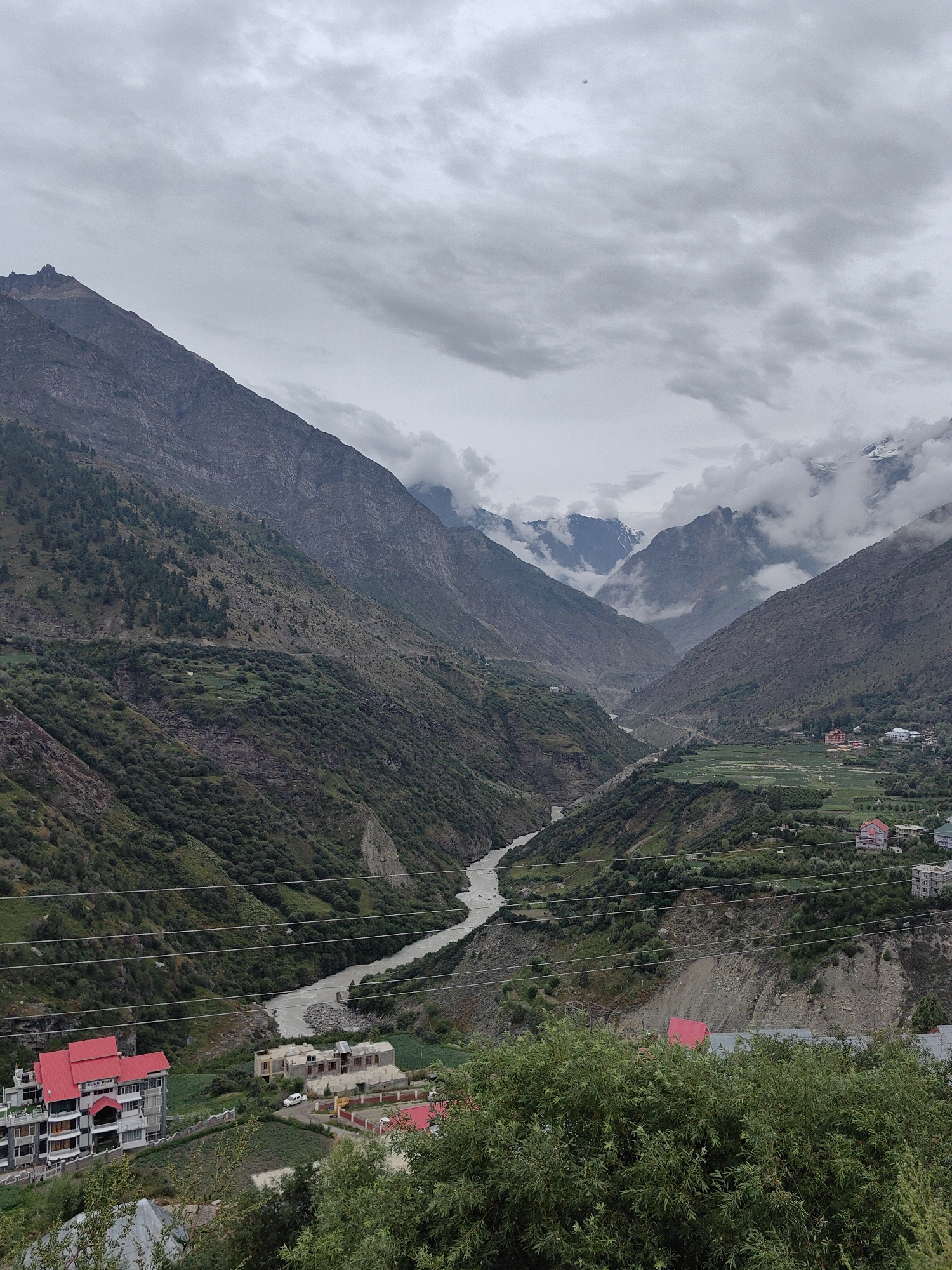 A high-angle, wide shot captures a cloudy, overcast day in the mountainous region of Keylang, India. The scene is dominated by a deep valley with a winding river flowing through its center. Steep, verdant mountainsides rise on either side of the valley, dotted with sparse trees and small patches of cultivation. In the foreground, a cluster of buildings with red roofs is visible, indicating a human settlement within the natural landscape. Power lines stretch across the frame, a common feature in such remote areas. The atmosphere is serene and somewhat dramatic, with the clouds adding a sense of depth and mystery to the rugged terrain.