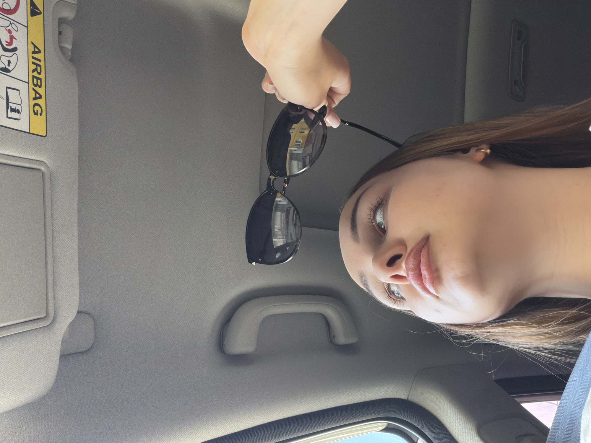 The image is taken from inside a car, looking upwards. A woman holds sunglasses in her hand. The scene includes the car's interior: a sun visor with an "AIRBAG" label, a grab handle, and a view of the sky through the windshield. The reflection in the sunglasses suggests an exterior environment, potentially a building facade, hinting at an urban area. There is no verifiable evidence to indicate it is Chaikovsky, Russia.