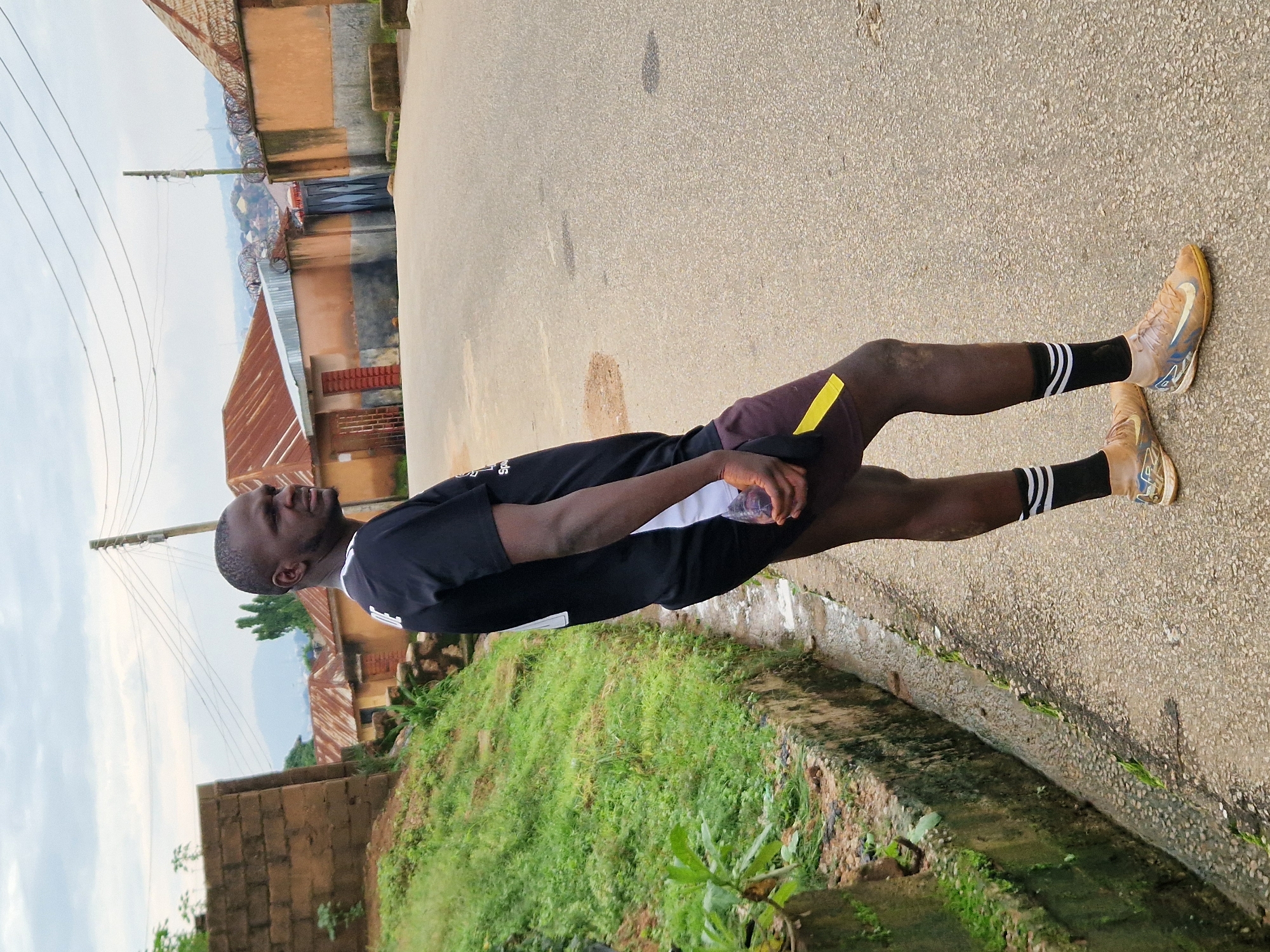 A man stands on an asphalt road in Kabong, Nigeria, looking to his right. He is wearing a black t-shirt with white accents, maroon shorts with a yellow stripe, black socks with white stripes, and tan athletic shoes. He is holding a clear plastic bottle in his right hand. To his left, there is a grassy embankment with a concrete drainage ditch. In the background, there are buildings with corrugated metal roofs and a distant view of hills. The sky is overcast.