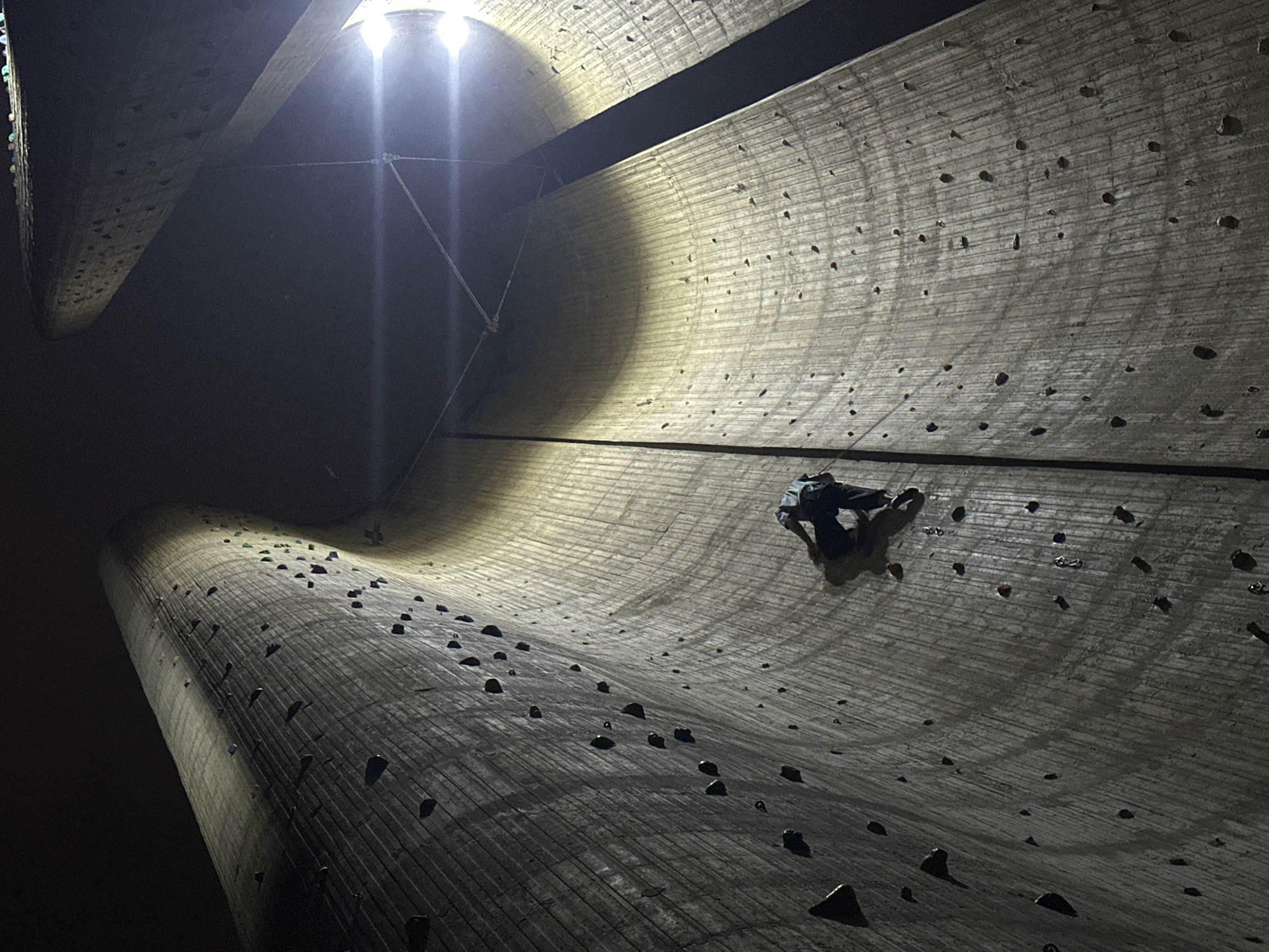 The photograph depicts a person rock climbing on a large, curved wall, likely a climbing structure, under bright artificial lighting. The wall is textured and equipped with numerous climbing holds. The climber is positioned on the right side of the frame, mid-climb. The environment is dark beyond the illuminated wall, suggesting nighttime. Location: Unknown, Unknown.