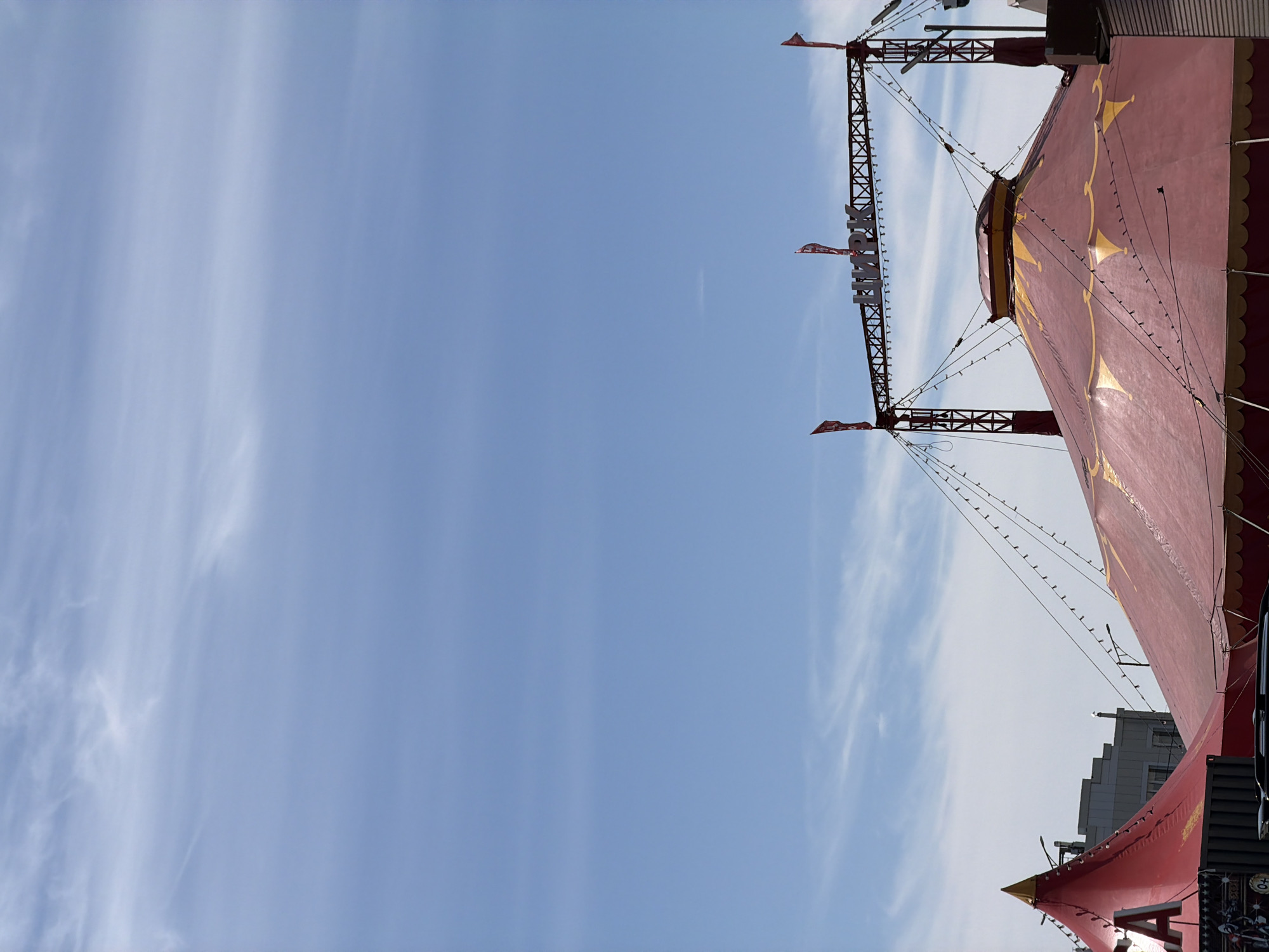 This is a low-angle, vertically oriented photograph of a circus tent under a bright blue sky with thin, wispy clouds. The tent is primarily red with gold decorative elements, including a patterned trim along its edges and star-like shapes on the fabric. Structural supports and rigging, likely part of the tent's construction or a supporting tower, are visible on the right side of the frame. Small flags and strings of lights are attached to these structures. Text is visible on one of the structural supports, appearing to read "ЦИРК" (Tsirk), the Russian word for "circus." Buildings are visible in the background on the far right, suggesting an urban or semi-urban setting. The lighting indicates it is daytime, likely midday, with a clear and pleasant weather day. No people are visible in the frame. The overall mood is festive and inviting, characteristic of a circus setting. The location context places this scene in Petropavlovsk-Kamchatsky, Russia.