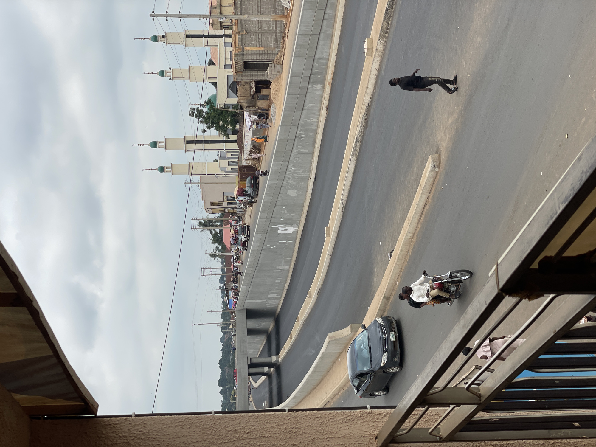 The image depicts a street scene in Akwanga, Nigeria, taken from an elevated perspective, possibly a balcony or high window. A multi-level road dominates the view, with a pedestrian walking on the upper level and a motorcycle carrying two people, as well as a dark-colored car on the lower level. In the background, a building with multiple minarets characteristic of a mosque is visible. Other buildings, vegetation, power lines, and people are also present along the road. The sky is overcast.