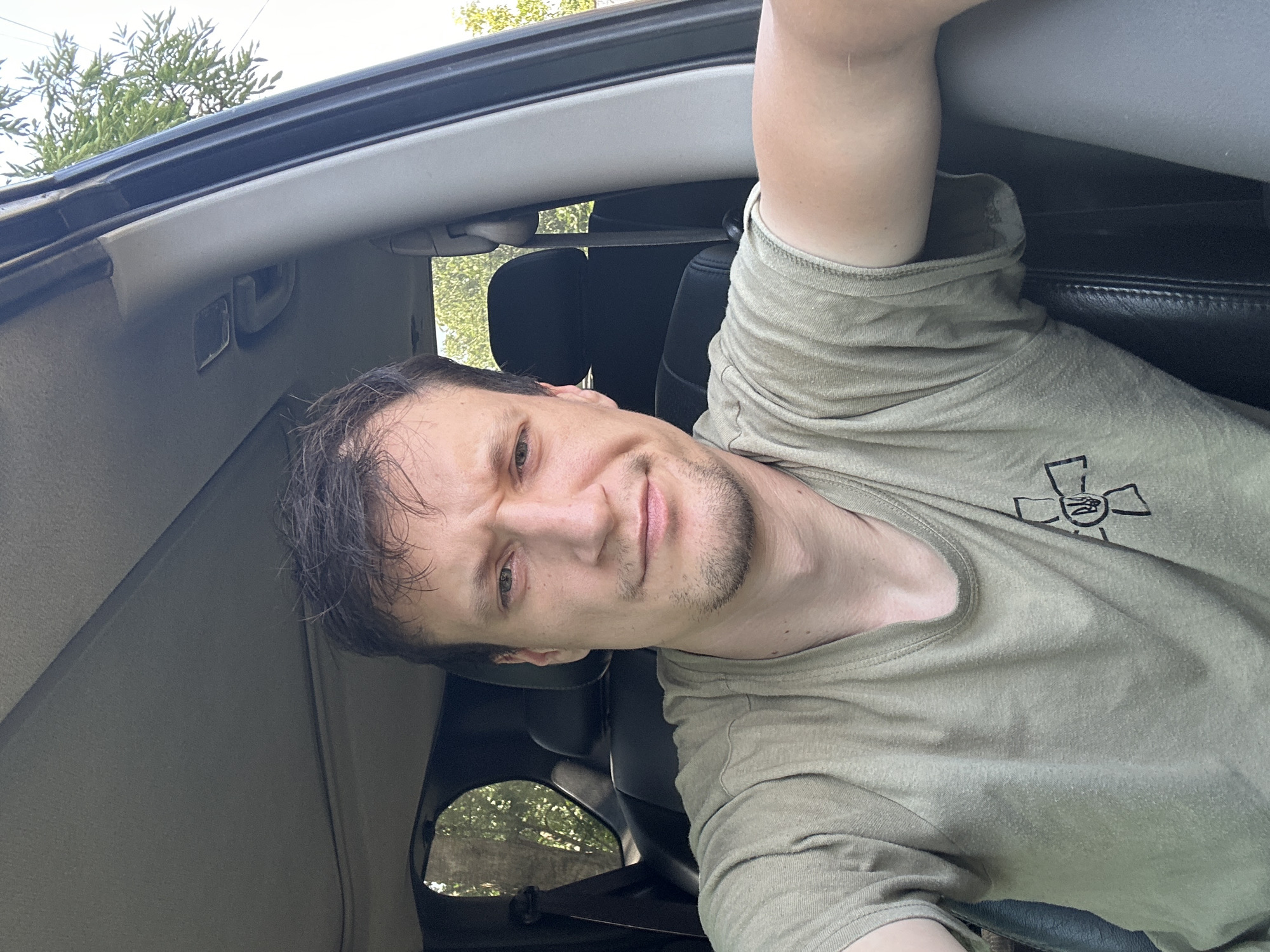 A close-up, frontal, slightly overhead, medium shot shows a fair-skinned man with short, dark brown hair, a light beard, and a slight frown. He is wearing a faded olive green t-shirt with a black emblem on the left chest featuring a stylized cross with a circular crest. He is seated inside a vehicle, facing the camera. The interior of the car is visible, including dark leather seats and a gray headliner. Through the open car window on the left, a blurred background of trees and foliage is visible. The right side of the image shows the interior pillar of the car and a bit of the exterior foliage. The lighting suggests it is daytime, with sunlight casting shadows. The image appears to be a selfie taken by the man. No specific details indicate the location being the city of Makiv, Ukraine.