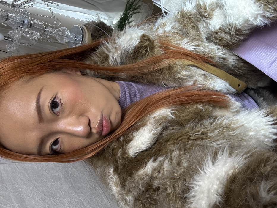 An Asian woman with reddish-brown hair lies on a white, textured surface. She wears a lilac-colored sweater and a brown and white furry coat. Her skin is dewy with blushed cheeks and glossy lips. A chandelier is visible in the upper left corner.