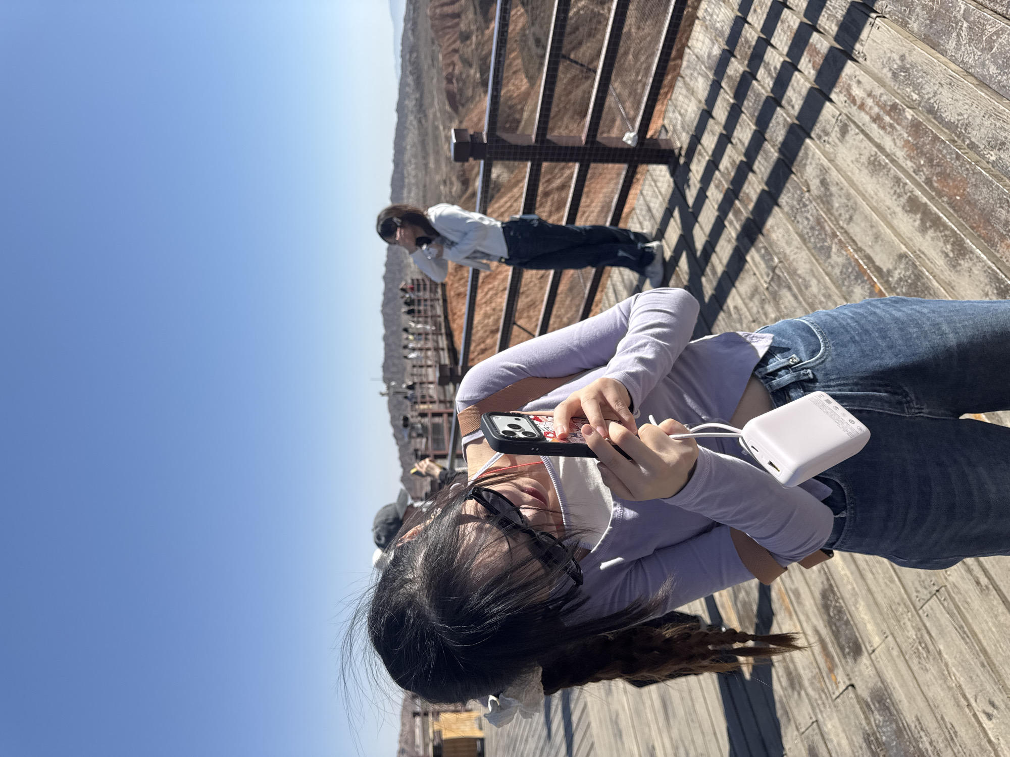 The image depicts an outdoor scenic viewpoint, likely located in Zhangye, China, given the distinctive reddish-brown geological formations characteristic of Danxia landforms. The scene is bathed in bright sunlight under a clear, vast blue sky, suggesting it is midday or early afternoon with excellent weather.
In the foreground, a young woman with long dark hair, wearing sunglasses on her head, a light purple long-sleeved top over a white undershirt, and blue jeans, is prominently featured. She is holding a black smartphone, likely an iPhone due to its camera array, with a red and white patterned case in both hands, appearing to be using it or capturing the view. A white, rectangular portable charger is connected to her phone by a white cable, hanging from her hands.
Further back on the wooden platform, another person is seen leaning against a dark railing, looking out into the distance at the expansive landscape. This individual wears a light-colored long-sleeved top and dark pants. In the blurred background, other indistinct figures are visible on what appears to be an extended viewing area.
The setting consists of a weathered wooden platform or walkway with sturdy dark railings, designed for visitors to observe the panoramic geological formations. The overall activity is sightseeing and enjoying the natural scenery. There is no clearly legible text visible on any of the objects in the image, although faint, unreadable icons are present on the portable charger. The scene conveys a sense of exploration and appreciation for a striking natural wonder.