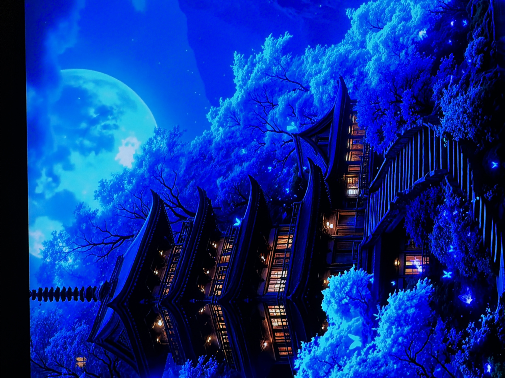 The image displays a vibrant and fantastical night scene dominated by deep and electric blue hues, set in what appears to be a magical forest or garden. A large, bright blue moon, partially veiled by soft blue clouds, illuminates the upper left portion of the sky, with faint stars twinkling around it. Below, a dense canopy of trees glows with an ethereal blue light, their foliage appearing iridescent and vibrant. Some trees display dark, intricate bare branches, contrasting with the glowing leaves. Scattered throughout the blue-lit trees are numerous glowing blue, butterfly-like or moth-like creatures, adding to the magical ambiance.
A dark, multi-storied building with distinctive, ornate, traditional East Asian architectural features, including peaked roofs and intricate details, occupies the central and right side of the frame. Warm, golden light spills from numerous windows, creating a striking contrast with the dominant cool blue tones of the environment. The building appears to be situated beside or above a calm body of water, as its detailed reflection is clearly visible below, mirroring its structure and the warm window lights. A bridge or walkway with vertical railings extends from the building into the blue-lit landscape. To the far left, a decorative, segmented chain-like element stretches horizontally, seemingly connected to the building. The scene, possibly depicting a fantastical rendition of nature and architecture in Sofiivska Borshchahivka, Ukraine, conveys a sense of enchanting mystery and serene wonder. There is no visible text or human activity.