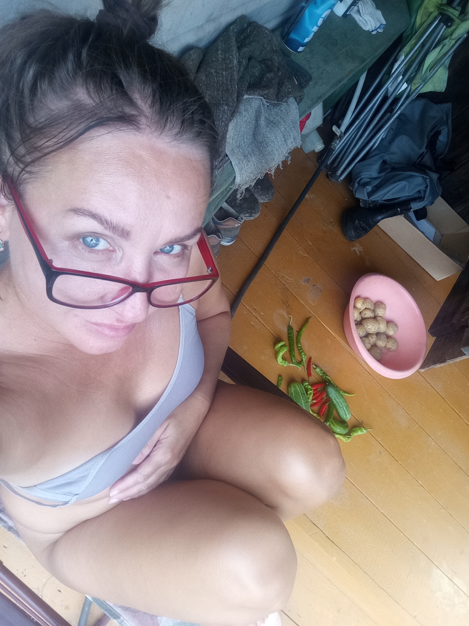 A high-angle, medium shot shows a woman with her hair in a bun, wearing glasses, and a light-colored bra. She is looking directly at the camera with blue eyes. Her hand is resting on her abdomen. The background is a wooden floor with various objects scattered around, including a pink bowl filled with potatoes, a pile of green and red chilies, and several small cucumbers. Also visible are a gray blanket, a black boot, a blue can, and metal poles. The scene appears to be indoors, likely a room, possibly in a residence located in the city of Novopokrovka, Russia.