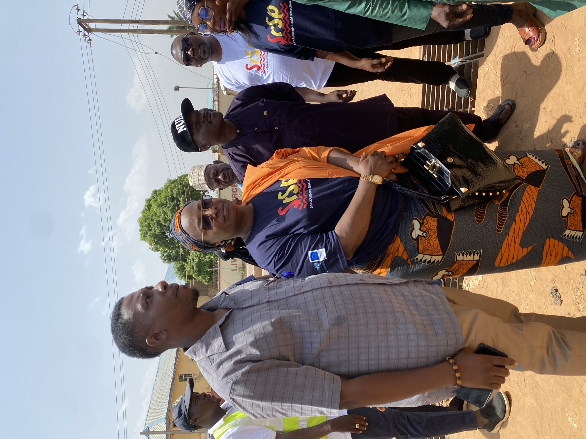 A group of people stand in Sabon Tasha, Nigeria, evident from the presence of power poles and overhead wires. The people, men and women, stand on bare ground. A woman in the center wears a navy blue top with “Shock-Responsive Social Protection” printed on it and carries a black, textured handbag. She also wears a patterned skirt. One man is wearing a hat with the word “NUIS” on it.