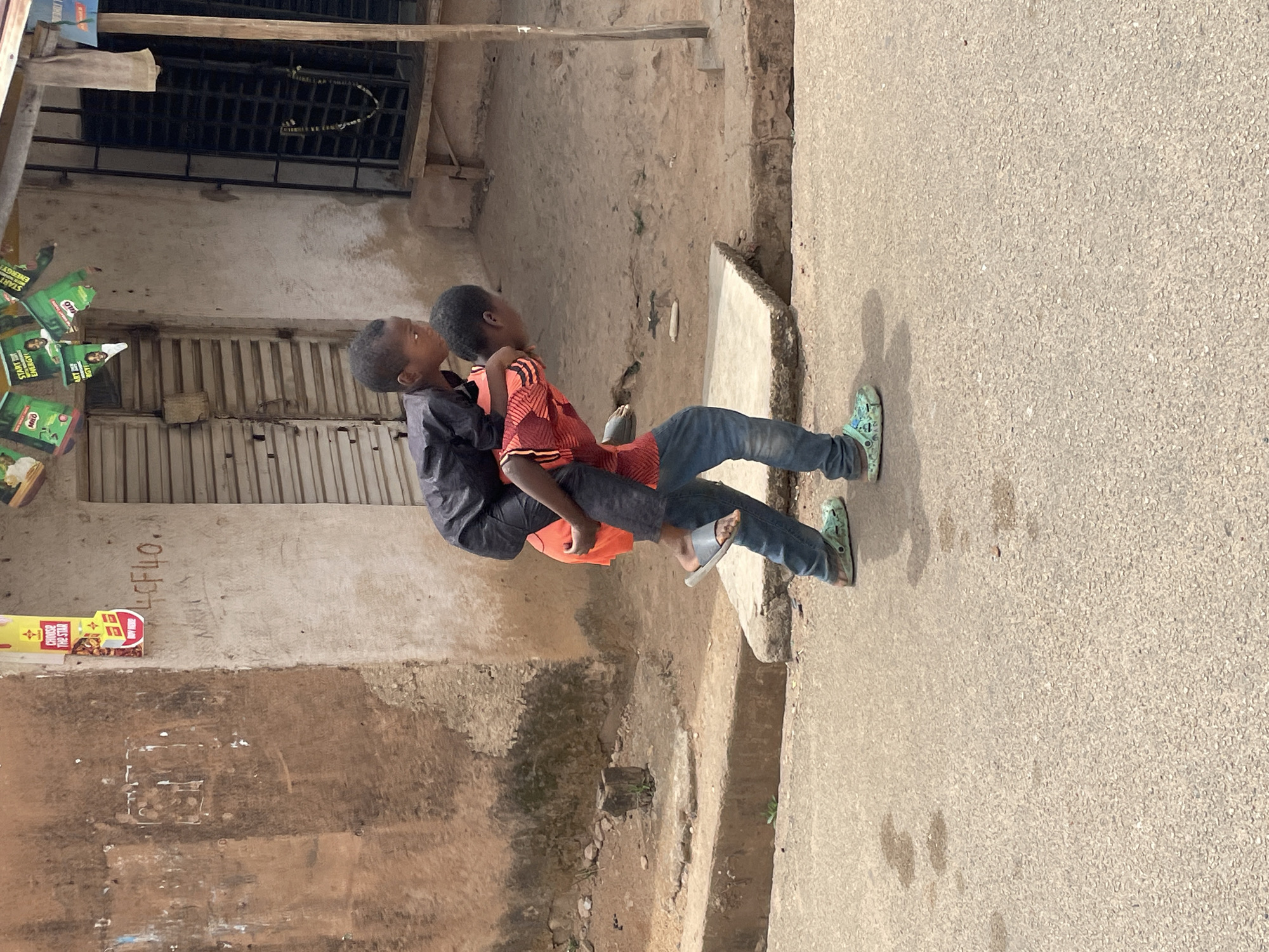 Two young boys are standing near a weathered building and a road in Jos, Nigeria. One boy is carrying the other on his back. The boy carrying has on a dark shirt and jeans. The boy being carried has on an orange shirt, jeans, and slides. The building has corrugated metal shutters and signage. The road is paved.