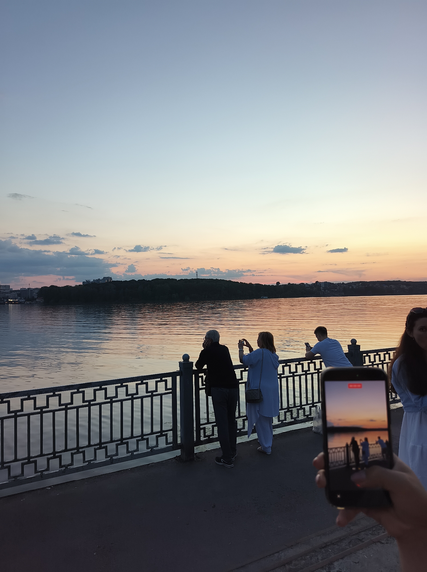 The image depicts a waterfront scene at sunset in Ternopil, Ukraine. Several people are standing along a promenade, which is separated from the water by a decorative metal railing. In the foreground, a person is holding a smartphone, capturing the view. The phone's screen displays a recording of the sunset, showing silhouettes of people against the water and sky. The sky is painted with hues of orange, pink, and blue, transitioning from the warm colors of the setting sun to the cooler tones of twilight. The water reflects the colors of the sky and the surrounding landscape. On the opposite bank of the body of water, a tree line is visible, with some buildings peeking through. The overall atmosphere is serene and tranquil, typical of an evening by the water.